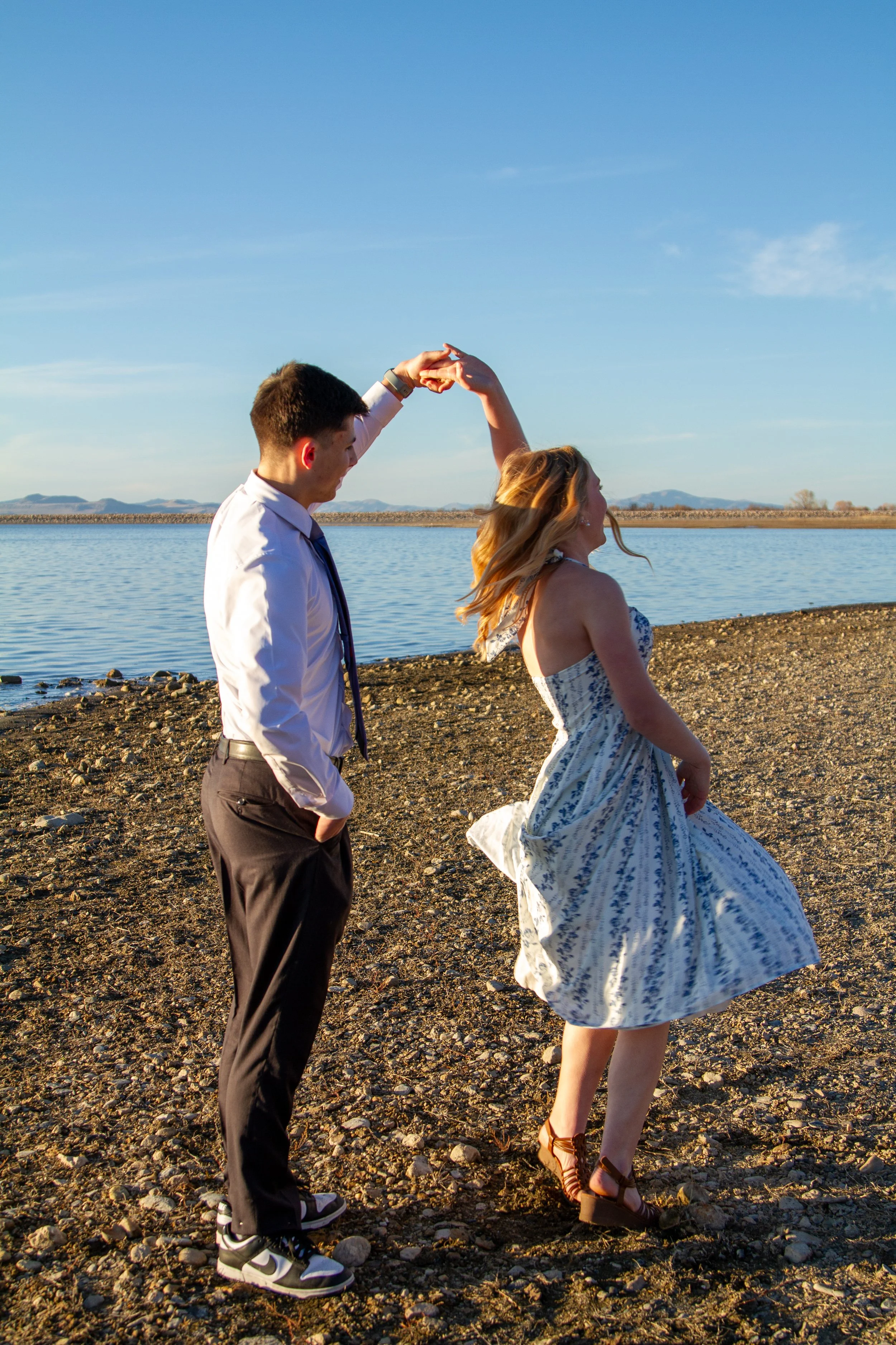 A young couple dancing by a body of water during the sunset. The man is wearing a white shirt, black pants, and sneakers. The woman is wearing a white patterned dress and brown shoes.