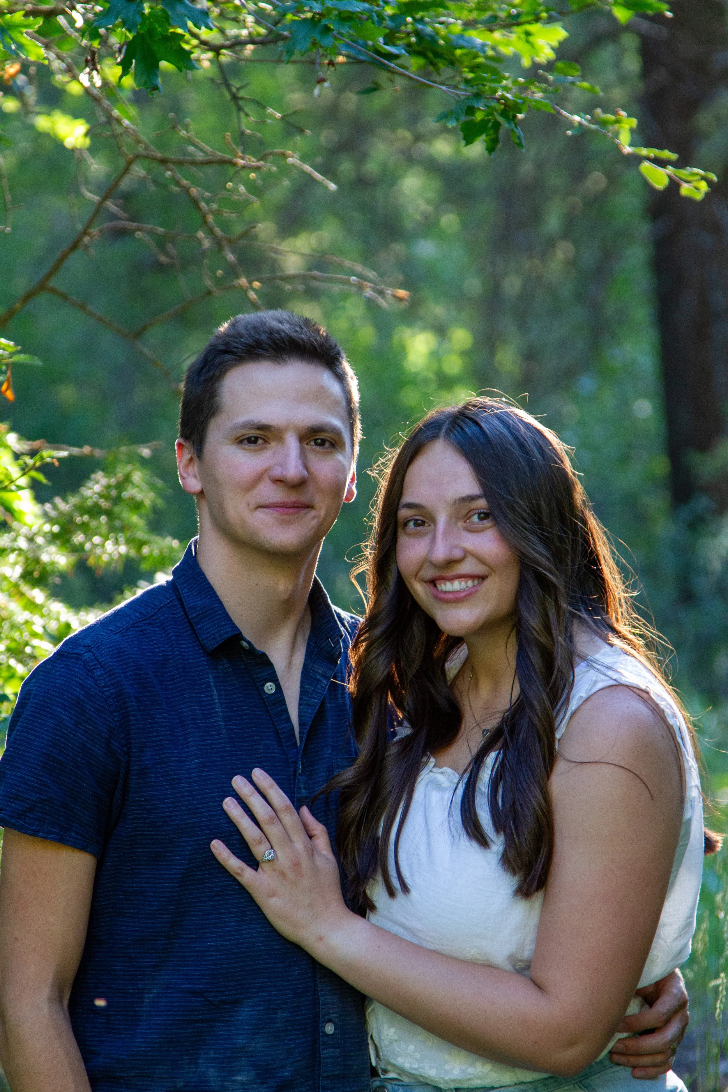 A young couple smiling outdoors with greenery and trees in the background. The woman has long dark hair and is wearing a light-colored sleeveless top, and the man has short dark hair and is wearing a dark blue shirt. They are standing close, with the