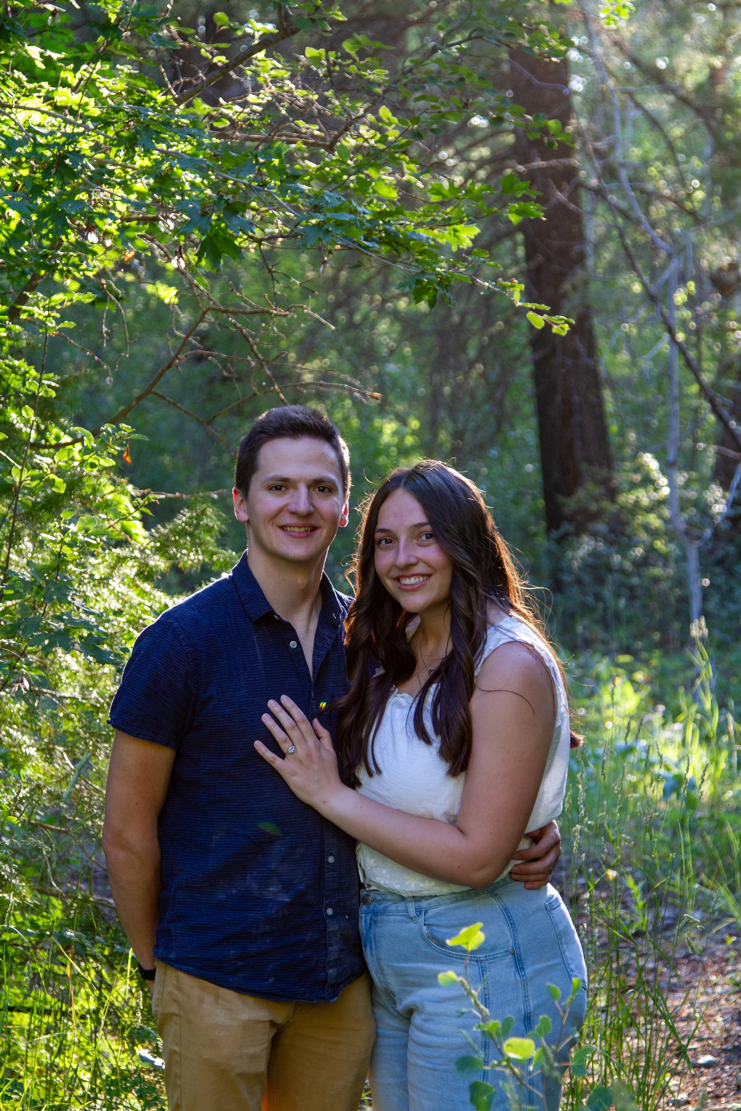 A young man and woman happily standing close together outdoors in a green forest, smiling at the camera. The man has short dark hair and is wearing a dark blue shirt and beige pants. The woman has long dark hair and is wearing a white sleeveless top 