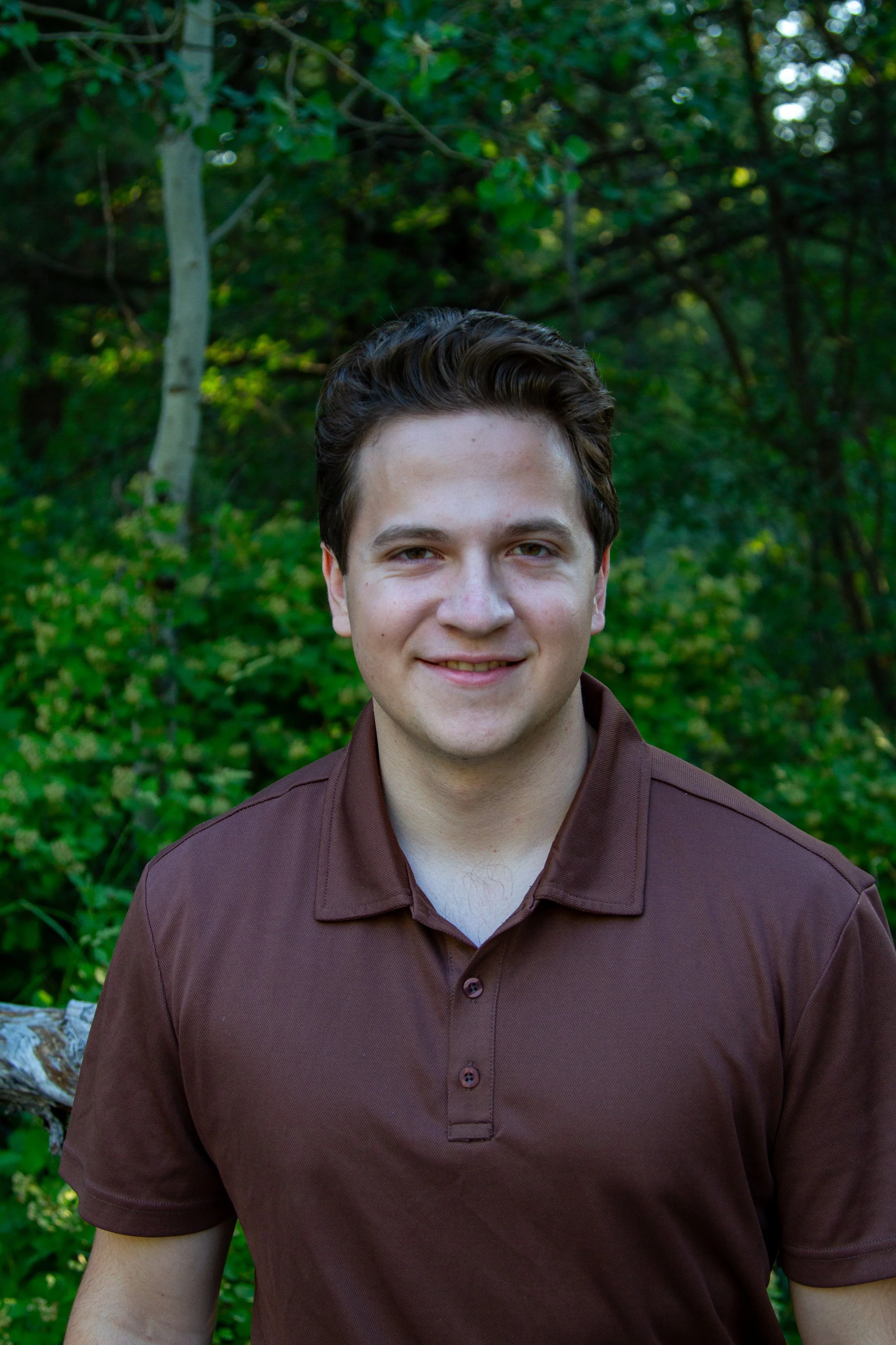 Young man with short brown hair smiling, wearing a brown polo shirt, outdoors with green trees and foliage in the background.