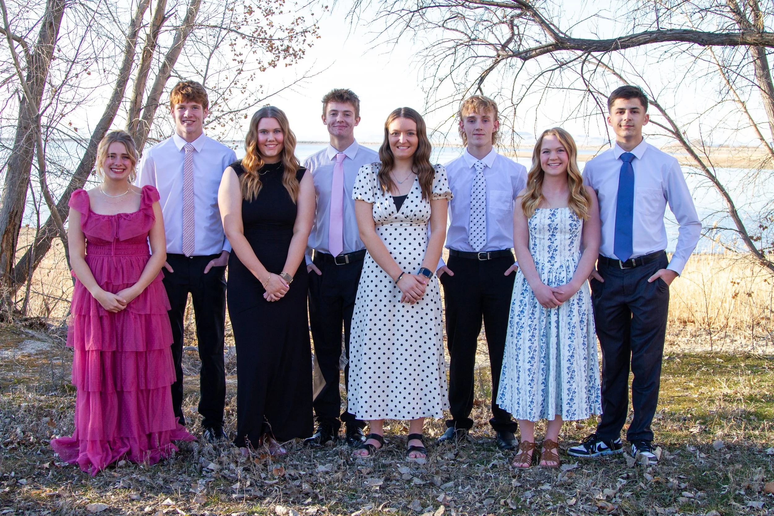 A group of nine young people dressed in semi-formal attire, standing outdoors on a grassy area with leafless trees and a body of water in the background.