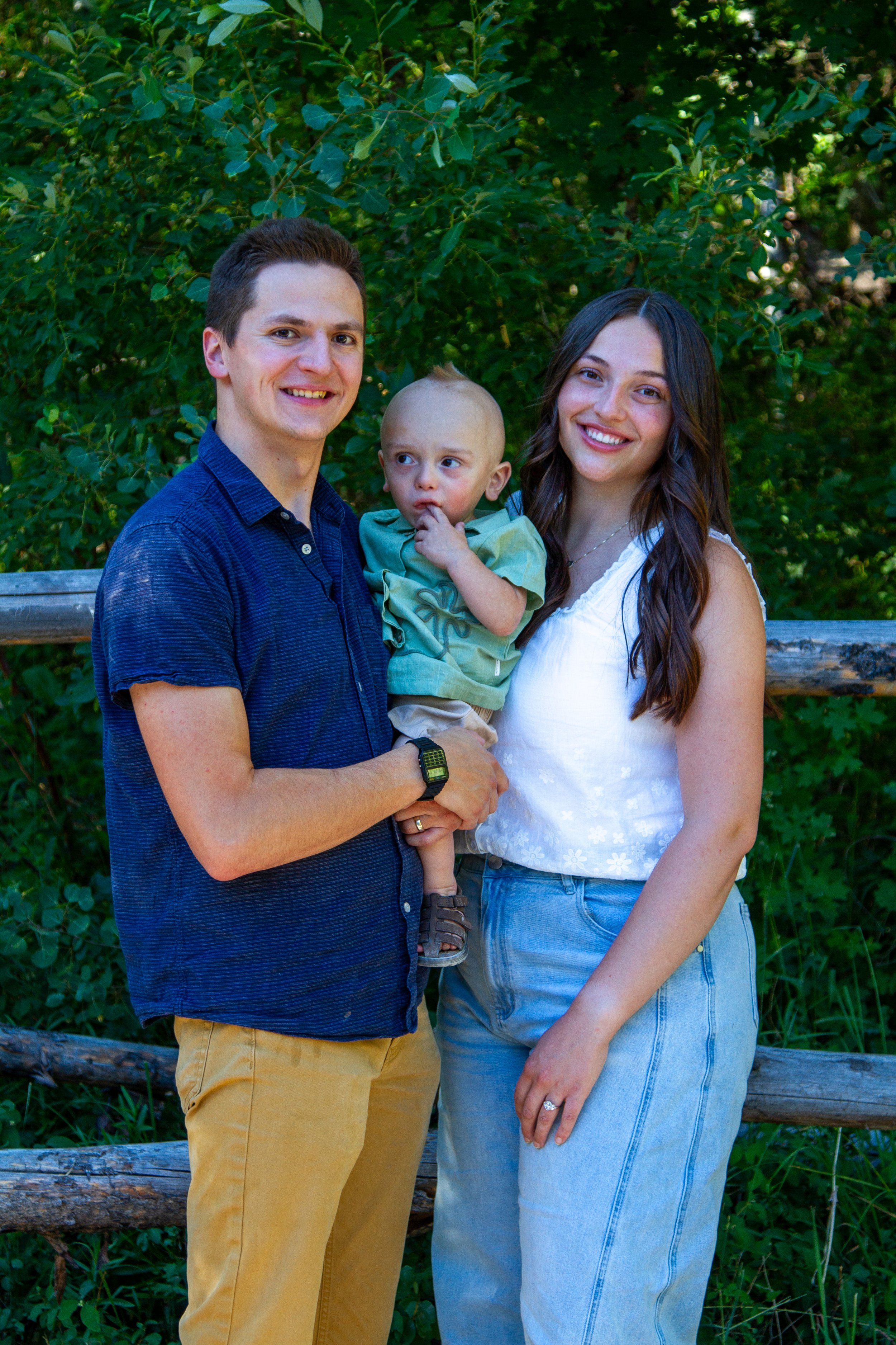 A family of three posing outdoors near a wooden fence with green foliage in the background. The man on the left wears a blue shirt and tan pants, the woman on the right wears a white sleeveless top and jeans, and the young child in the middle, held b