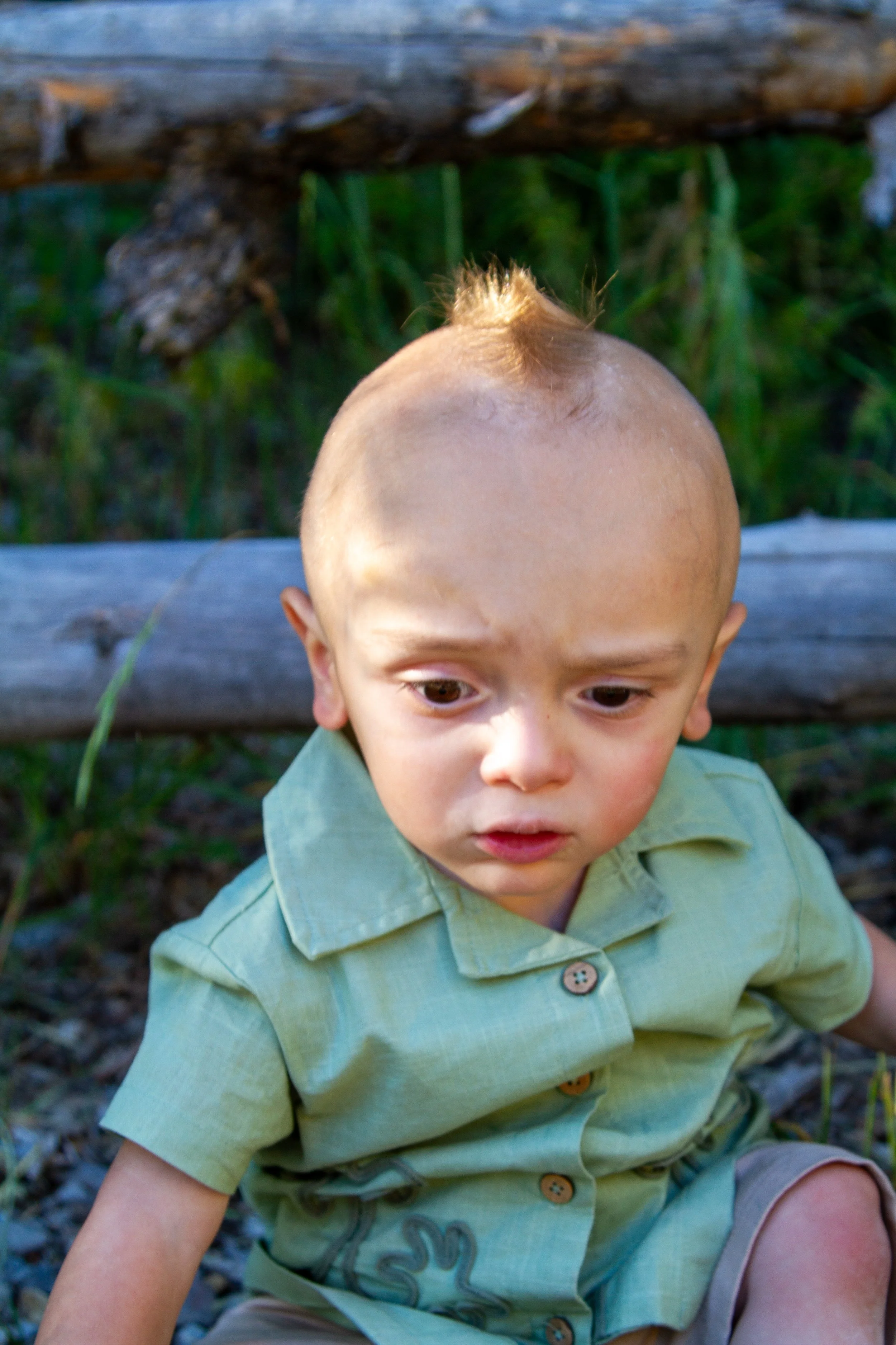 Young boy with a unique mohawk hairstyle, wearing a light green short-sleeved shirt with buttons, sitting outdoors on a bed of small stones with green grass in the background.