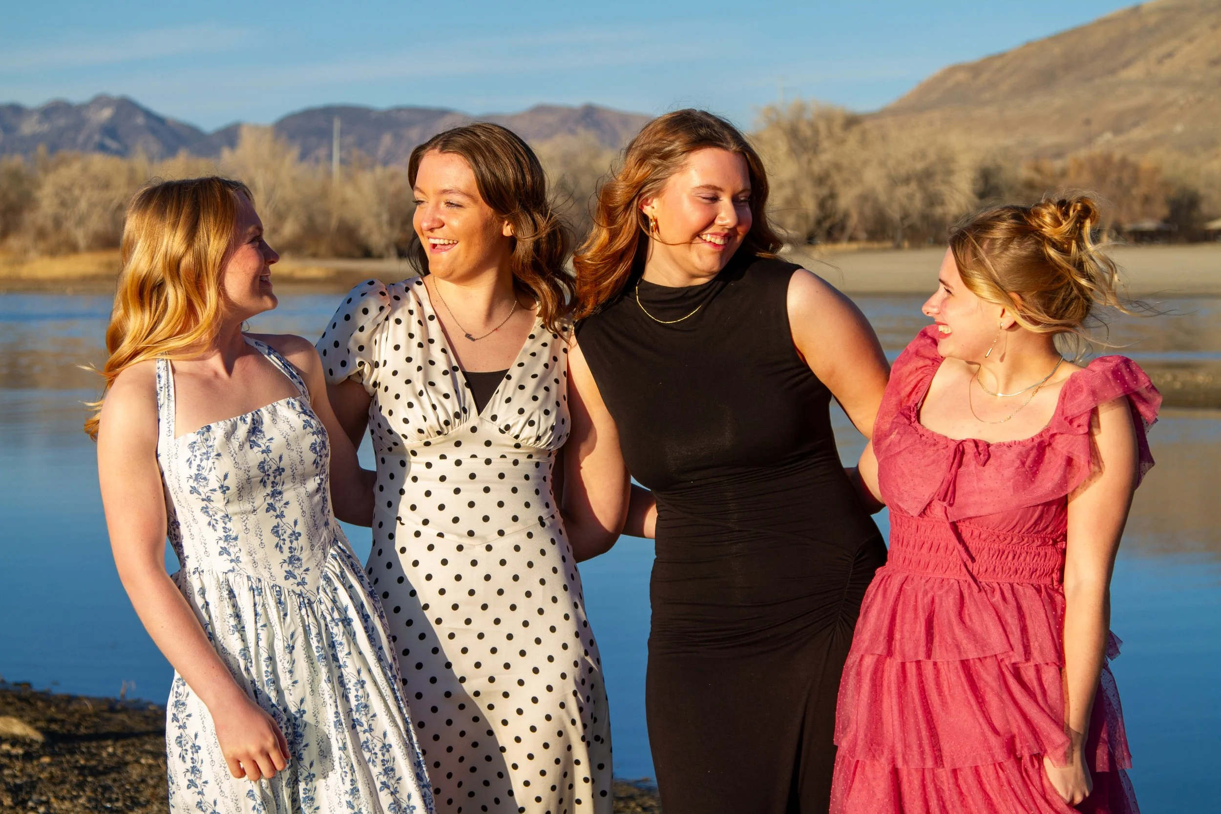 Four women smiling and walking together near a river with mountains in the background during sunset.