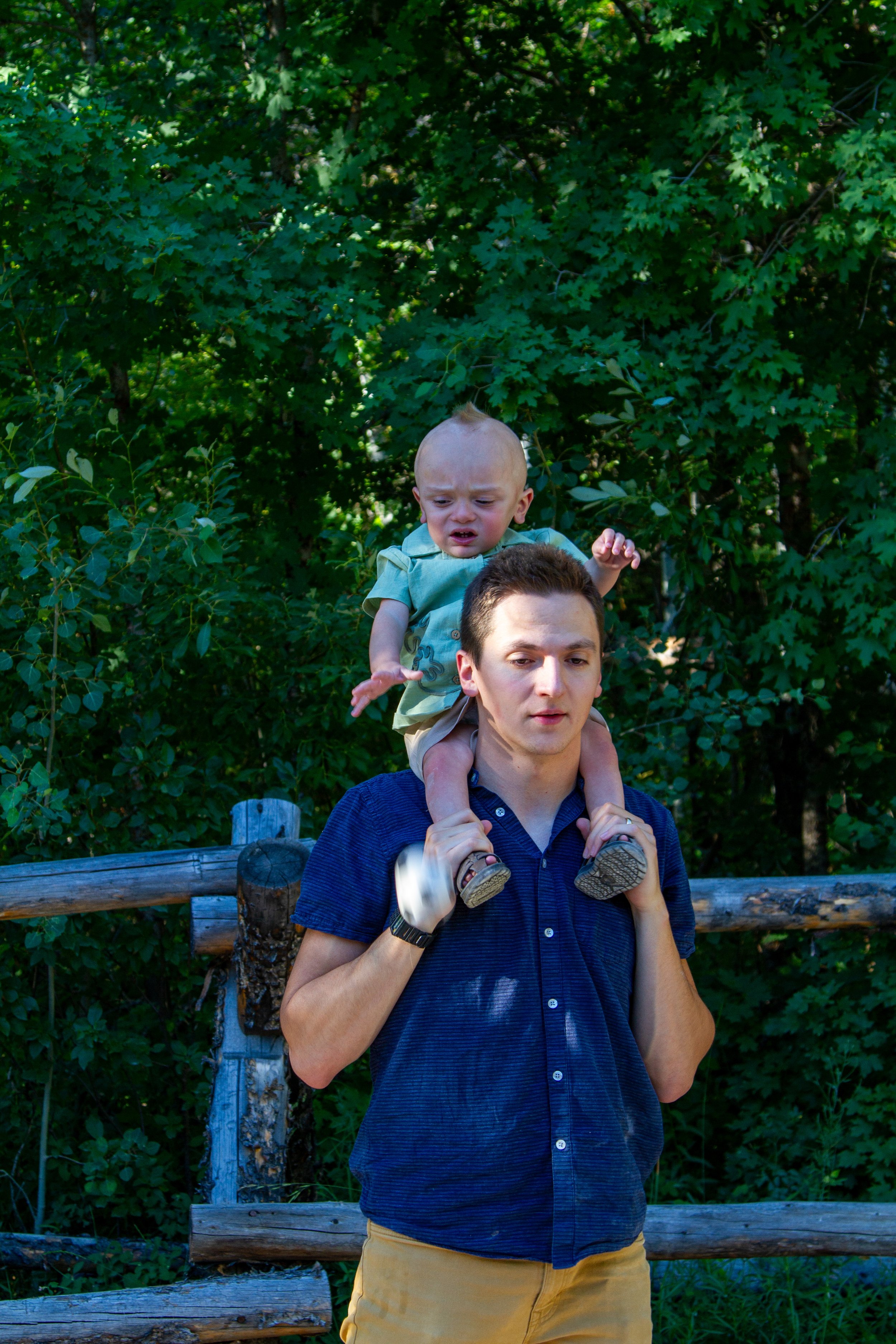 A man with a young child on his shoulders in front of green leafy trees. The adult man is wearing a dark blue short-sleeved shirt and yellow pants, and the child is wearing a light green shirt.