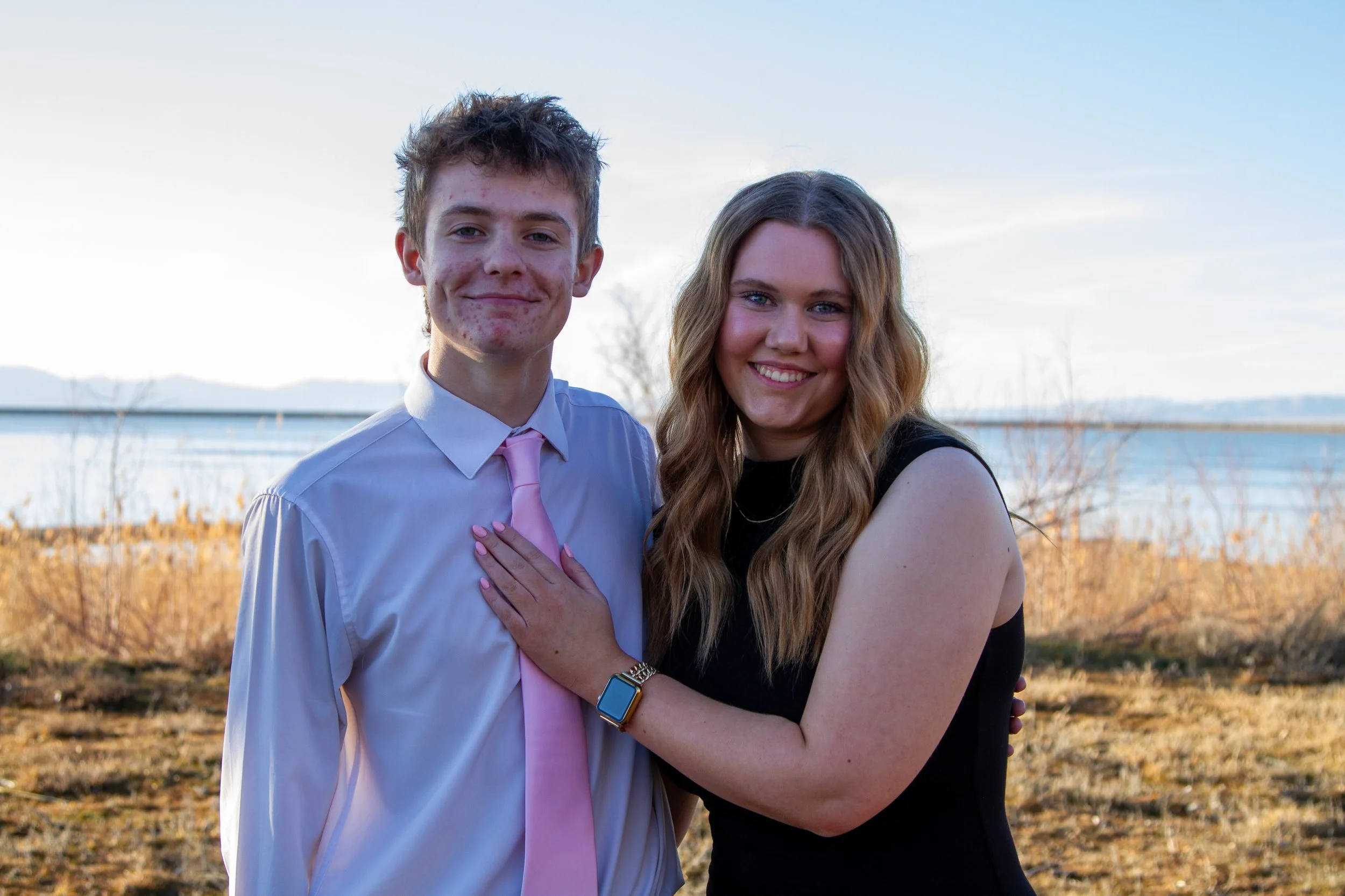 A young man and woman standing outdoors near a body of water on a clear day, smiling at the camera. The young man is wearing a white shirt with a pink tie, and the young woman is dressed in a black sleeveless top. She has her hand on his chest and we