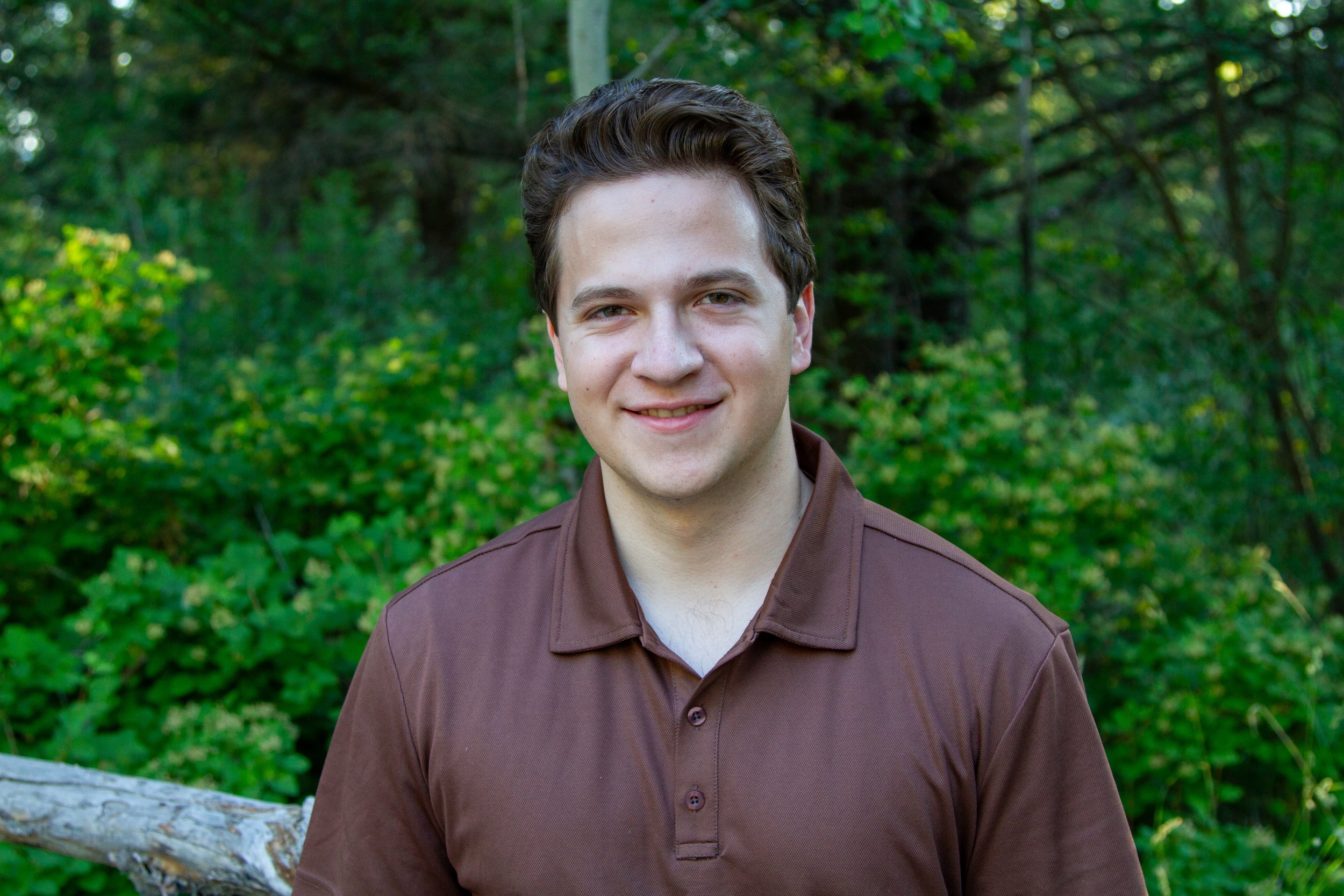 Young man with brown hair and a brown shirt standing outdoors in a lush green forest.