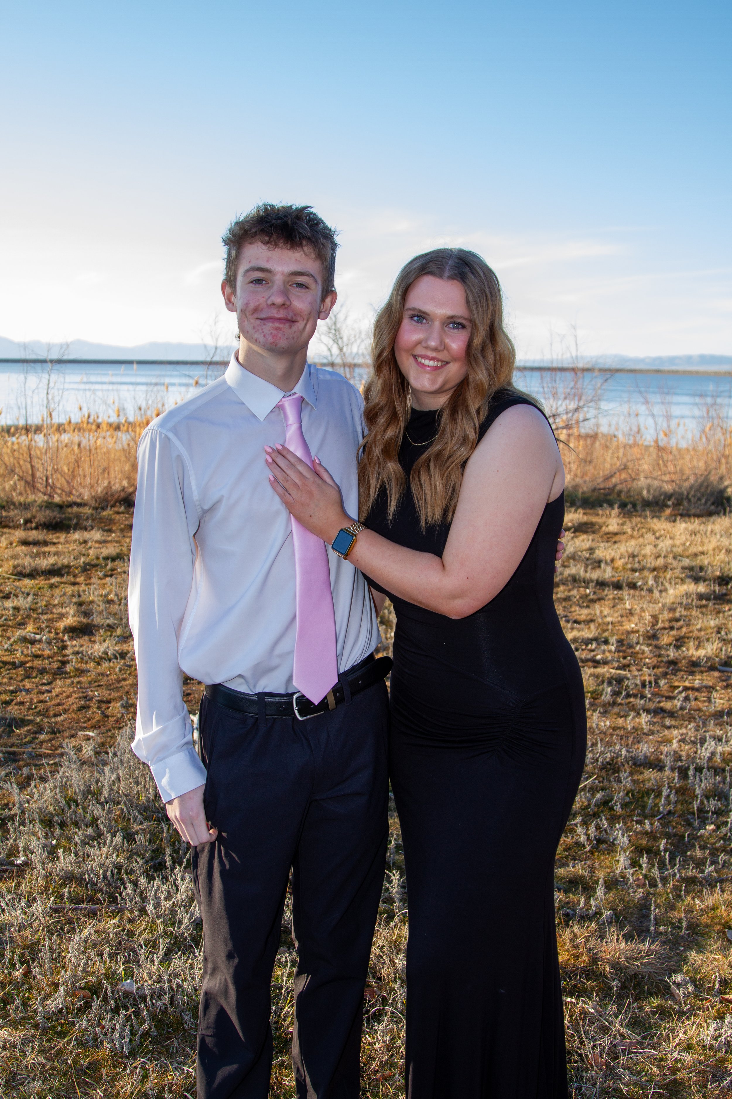 A young man and woman standing outdoors near a body of water, smiling at the camera. The man is wearing a white dress shirt, pink tie, and dark trousers. The woman is wearing a black sleeveless dress and has a smartwatch on her wrist. The background 