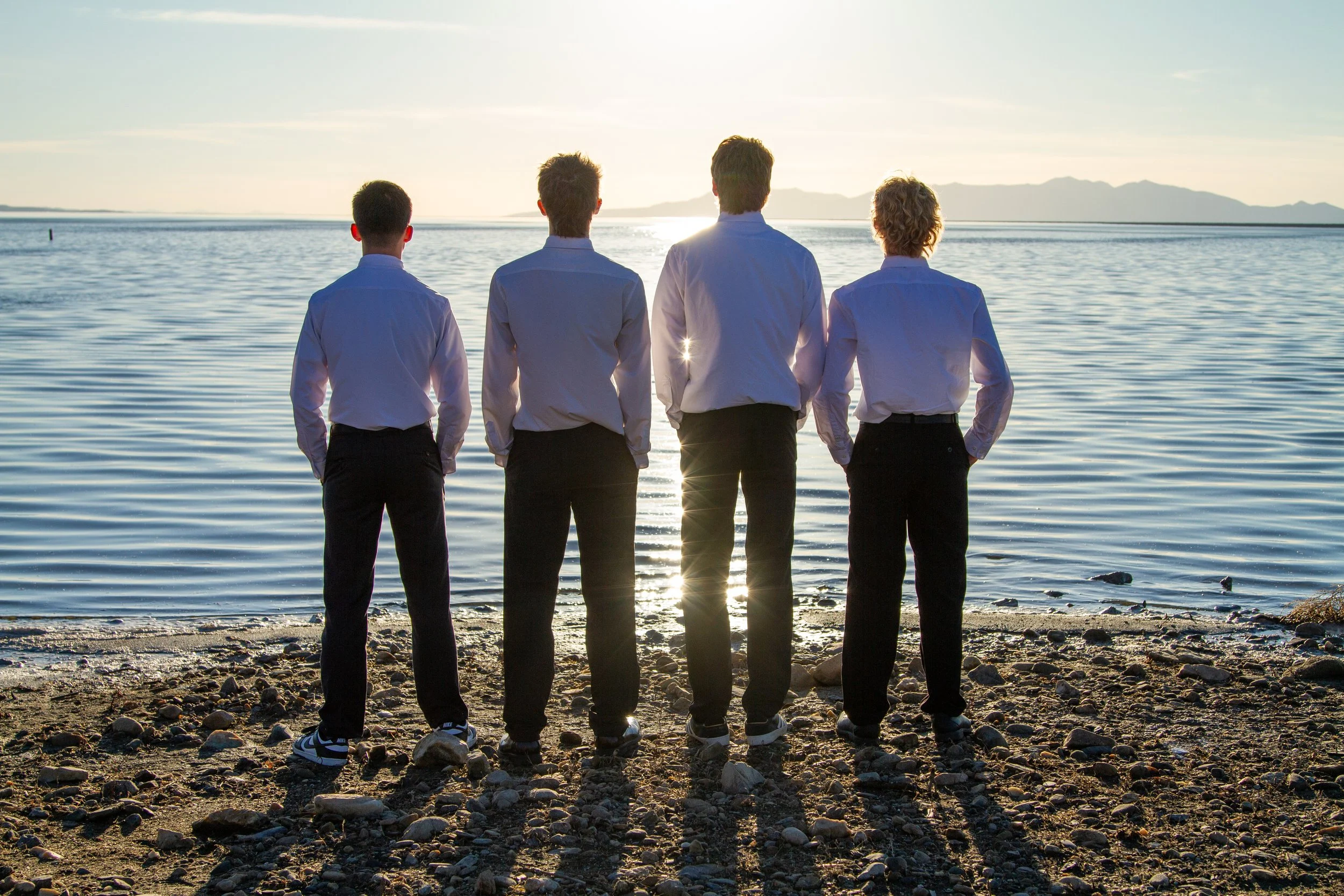 Four men dressed in white shirts and black pants stand on a rocky beach facing a calm lake during sunset.
