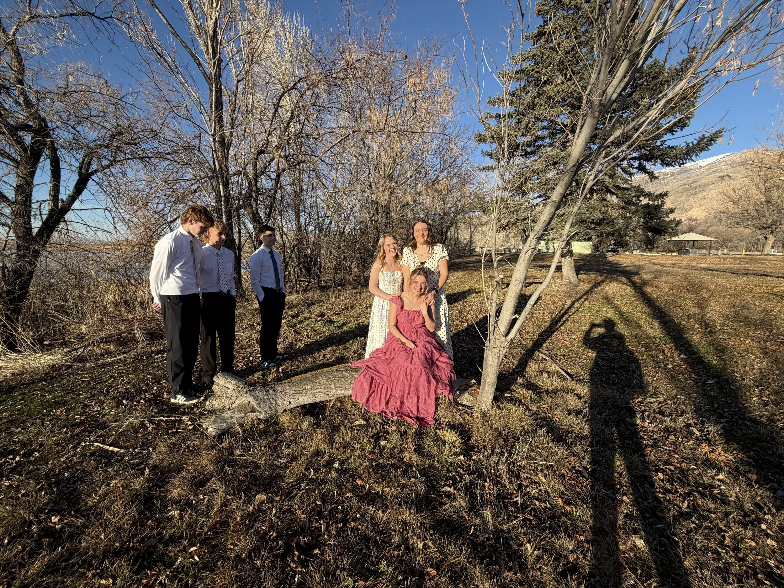 A group of six people, of which three are dressed in formal attire and three in casual dresses, are outdoors in a park-like setting with bare trees. They are standing and sitting around a fallen tree trunk, with mountains visible in the background on