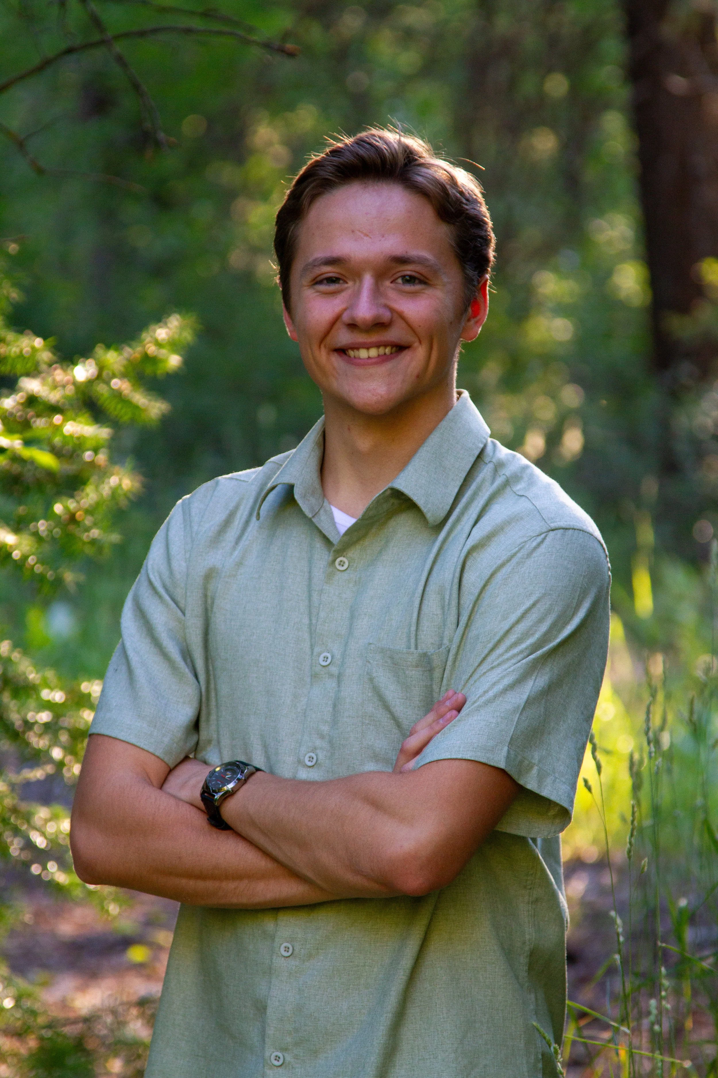 A young man with short brown hair and fair skin, smiling with arms crossed, standing outdoors in a wooded area with greenery and sunlight filtering through trees.