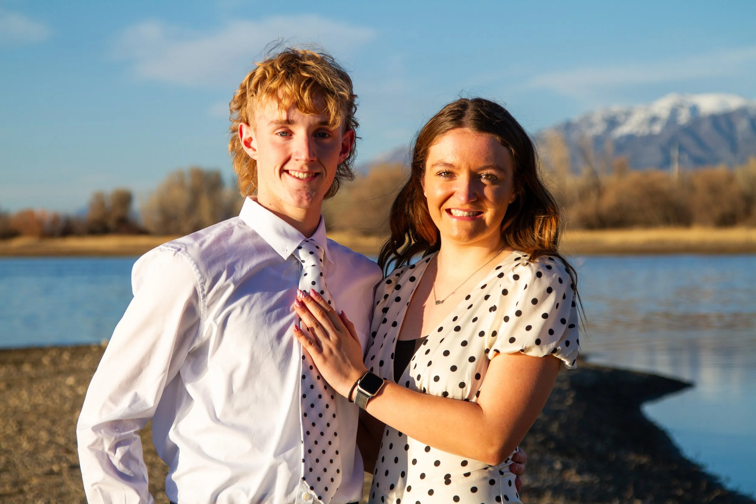 A smiling young man and woman standing close together outdoors near a lake, with mountains in the background, during sunset.