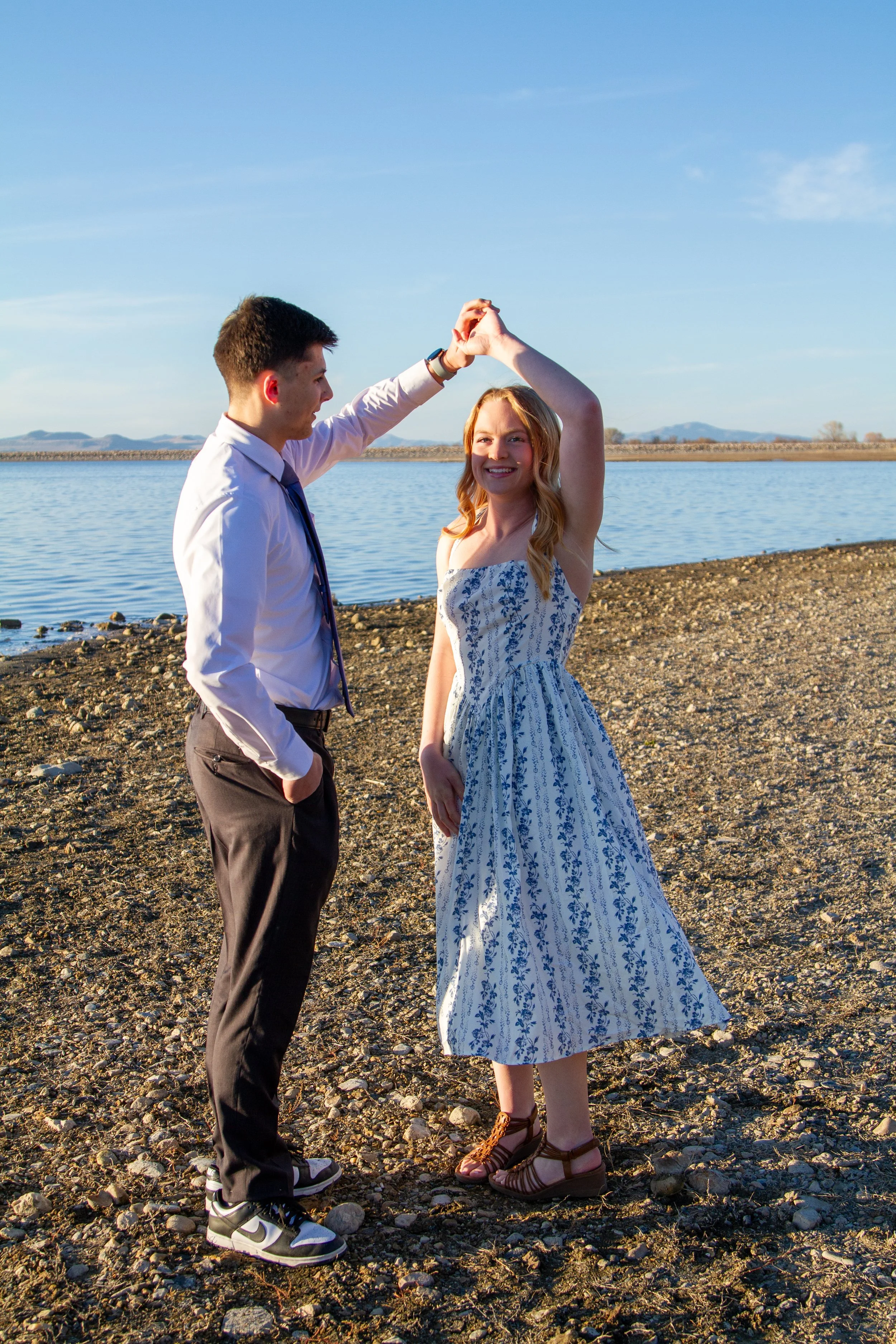 A young couple dancing on a rocky beach near a body of water under a blue sky.