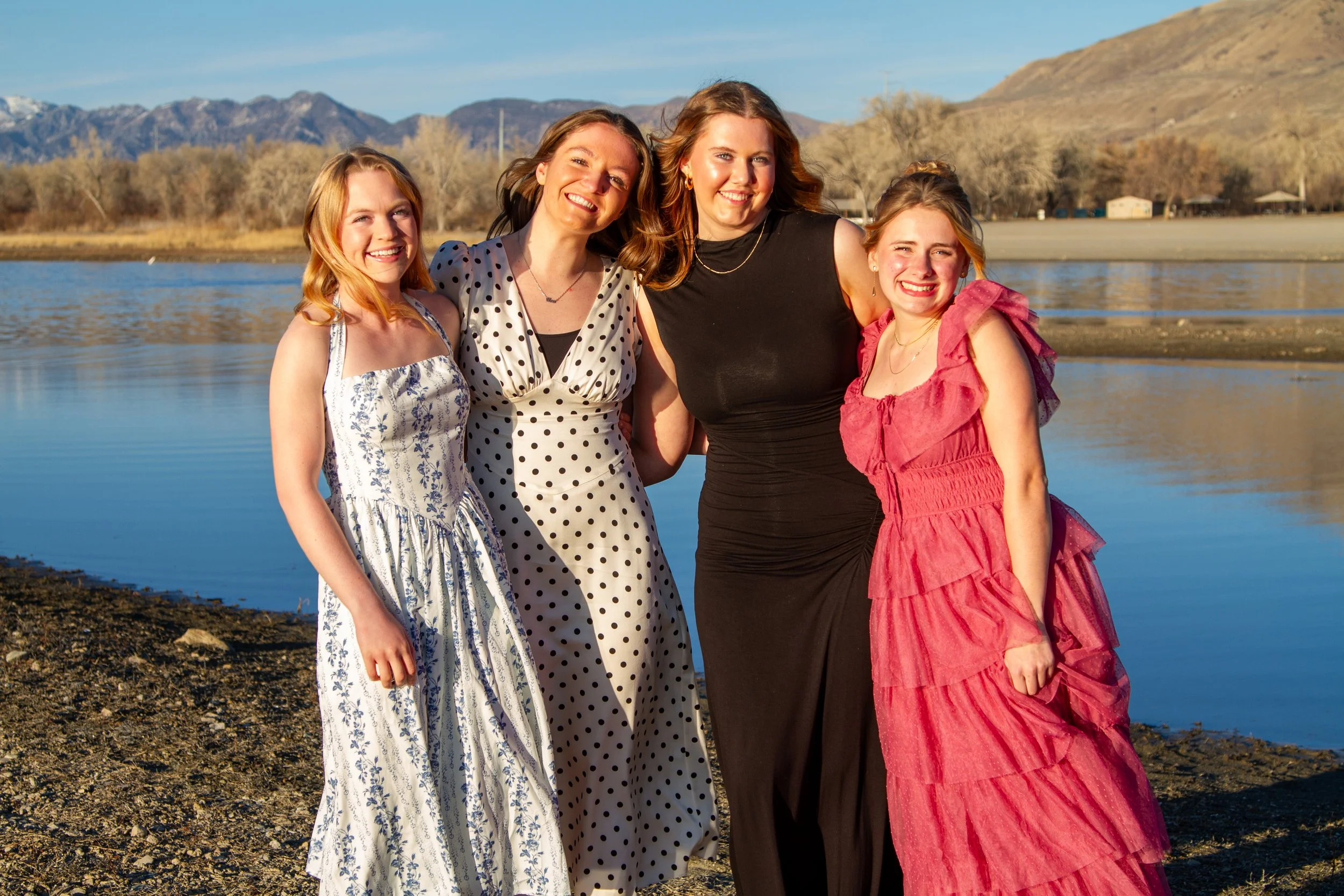 Four women standing by a lake on a sunny day, smiling and dressed in summer dresses, with mountains in the background.