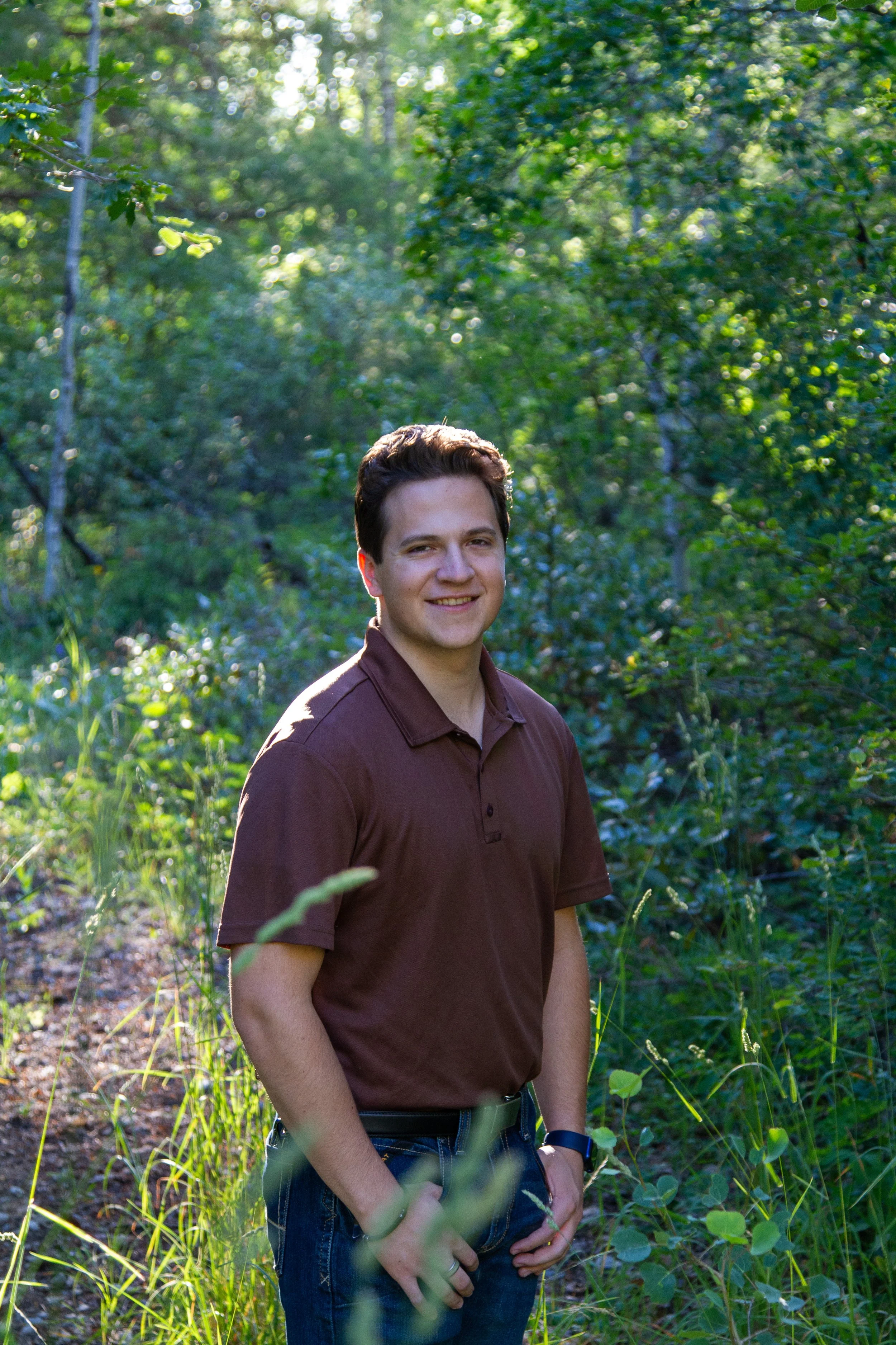 A young man with dark hair, smiling, standing in a green wooded area with sunlight filtering through the trees.