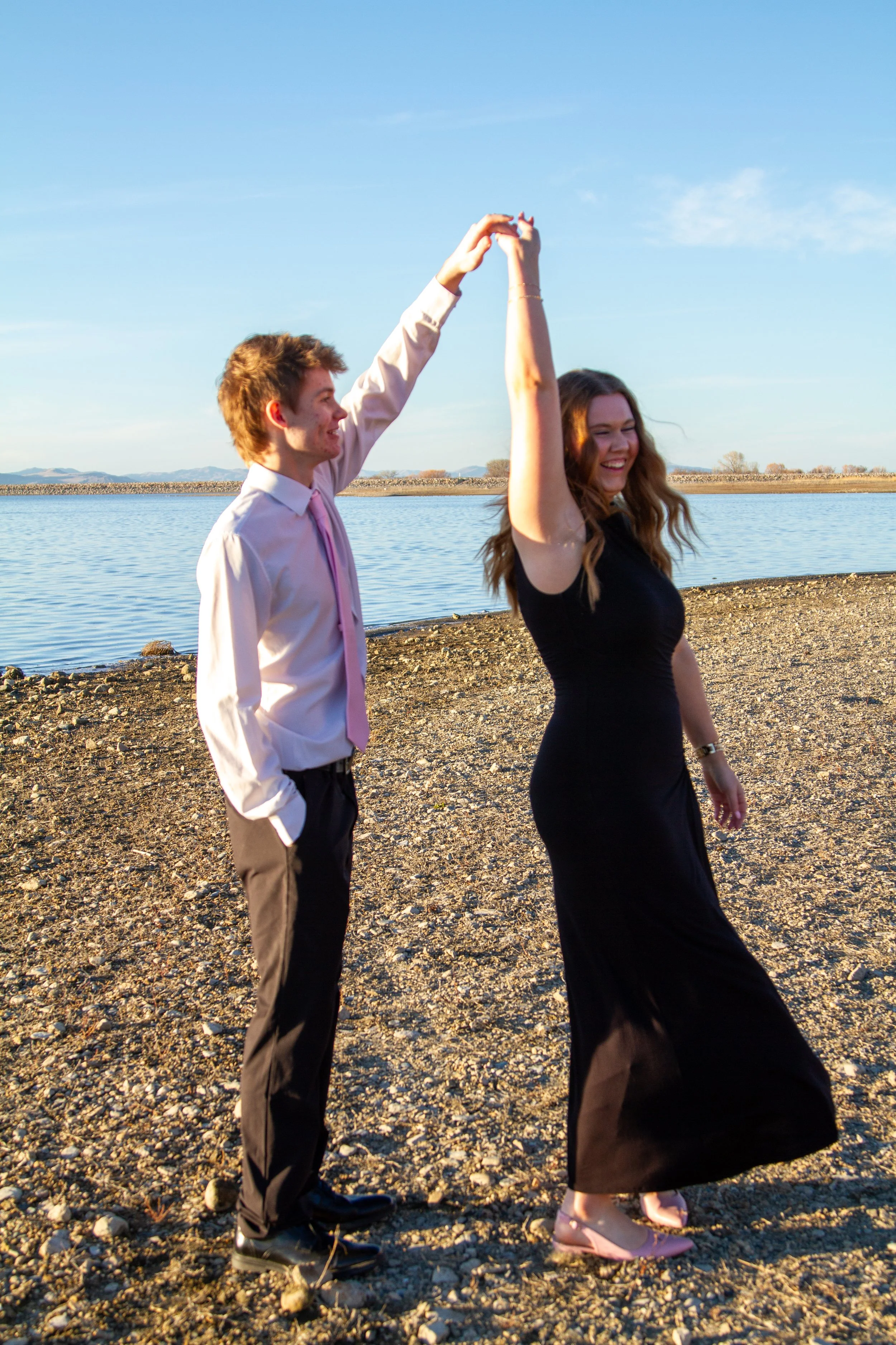 A young man and woman dancing by a lake on a sunny day, with the man twirling the woman as they both smile.