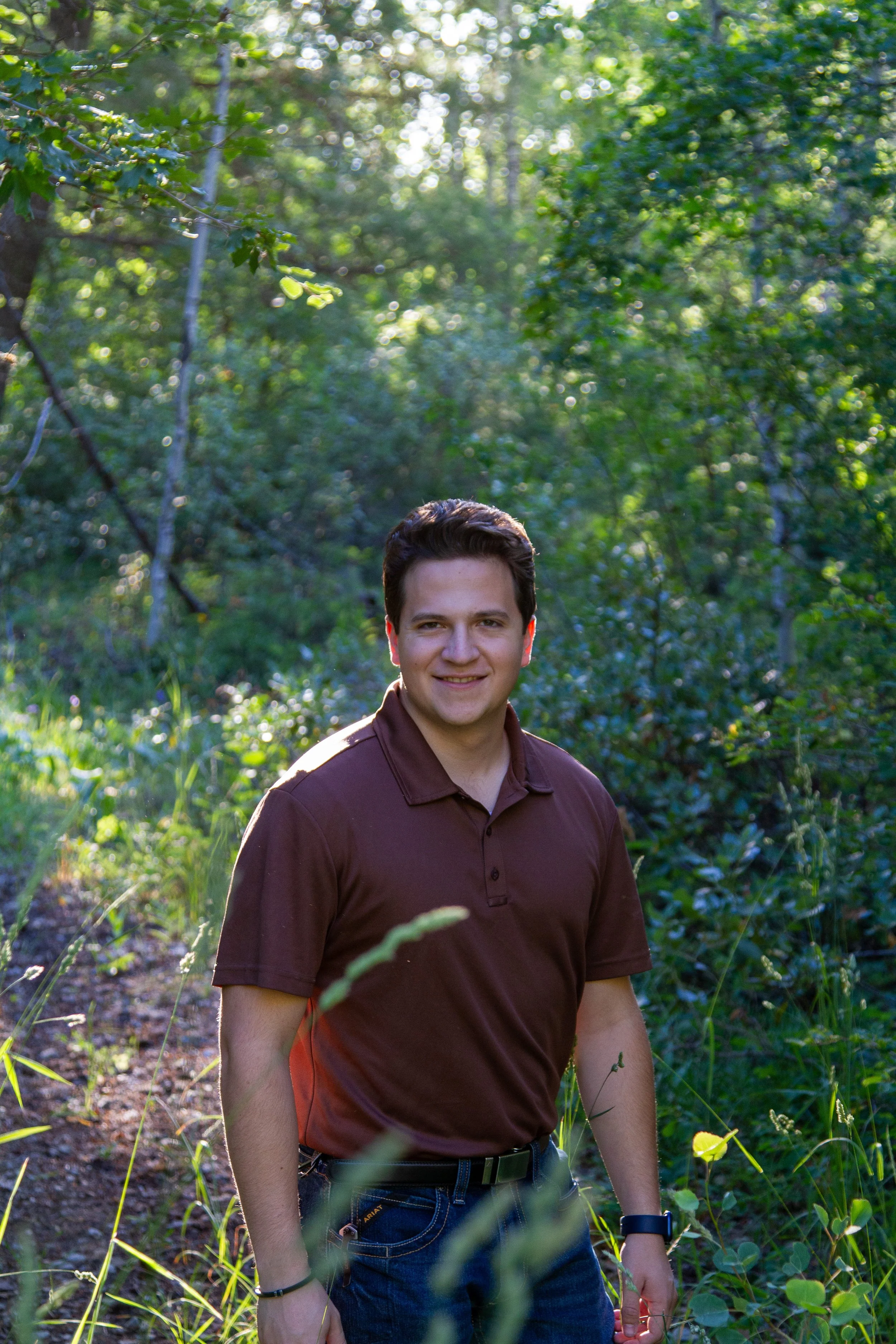 A young man with dark hair and a light complexion is standing outdoors in a lush, green forest during daytime. He is wearing a brown polo shirt, blue jeans, and a black watch on his left wrist. He is smiling and looking at the camera.