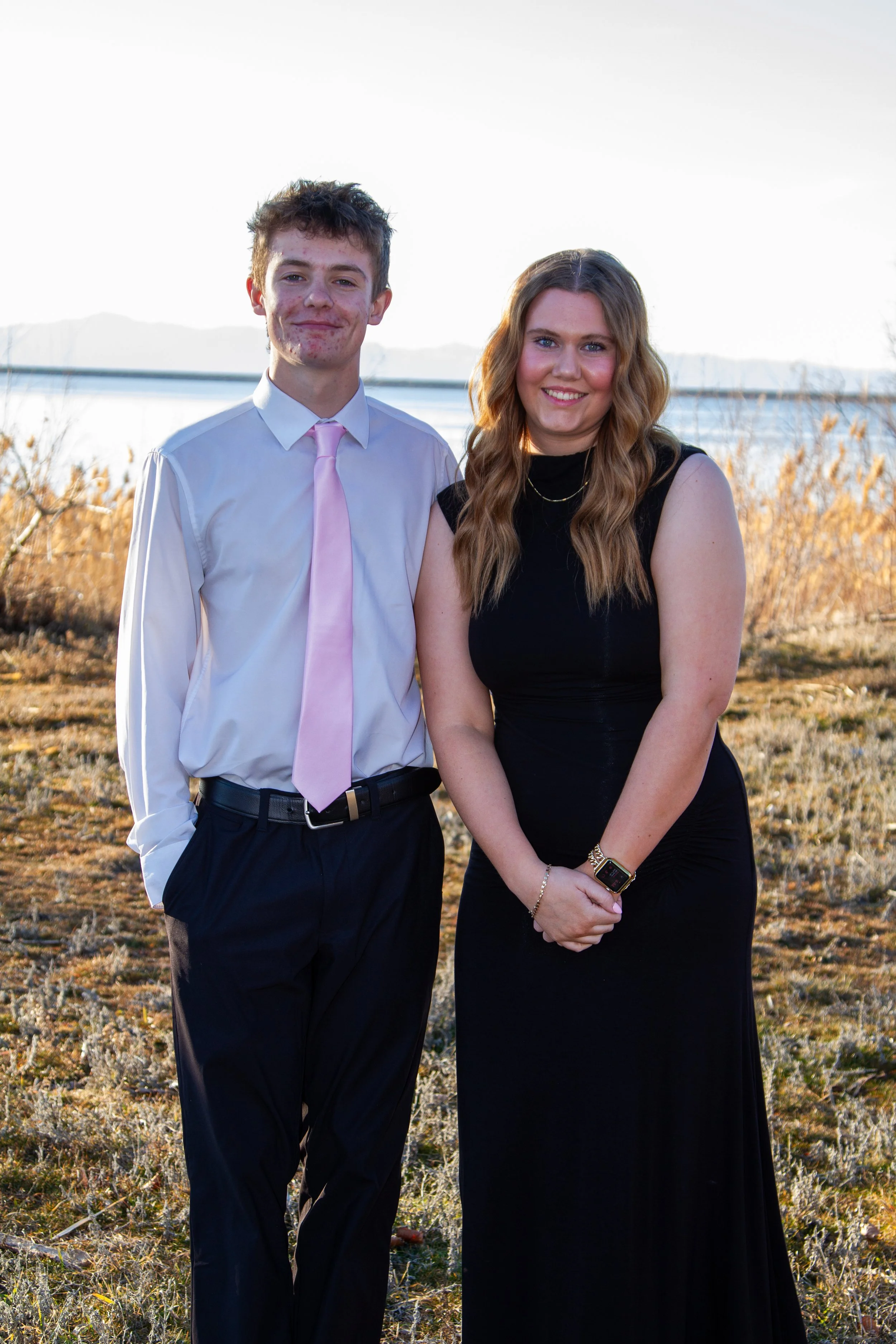 A young man and woman standing outdoors by a lake during sunset. The man is wearing a white shirt with a pink tie and dark pants. The woman is dressed in a black sleeveless dress, with long wavy hair, smiling at the camera.
