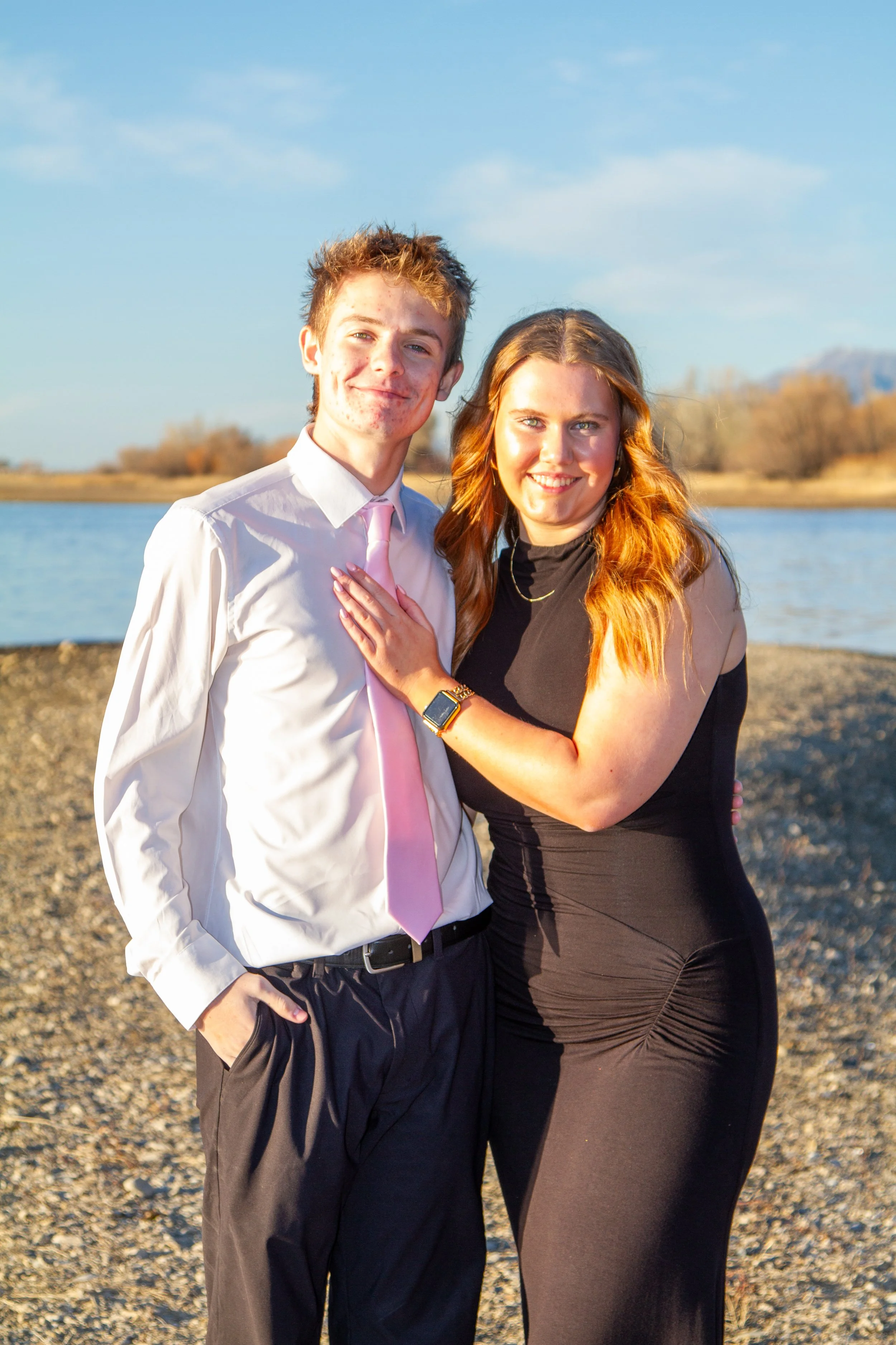 A young man in a white shirt and pink tie, and a woman in a black dress, standing together outdoors near a body of water during sunset.