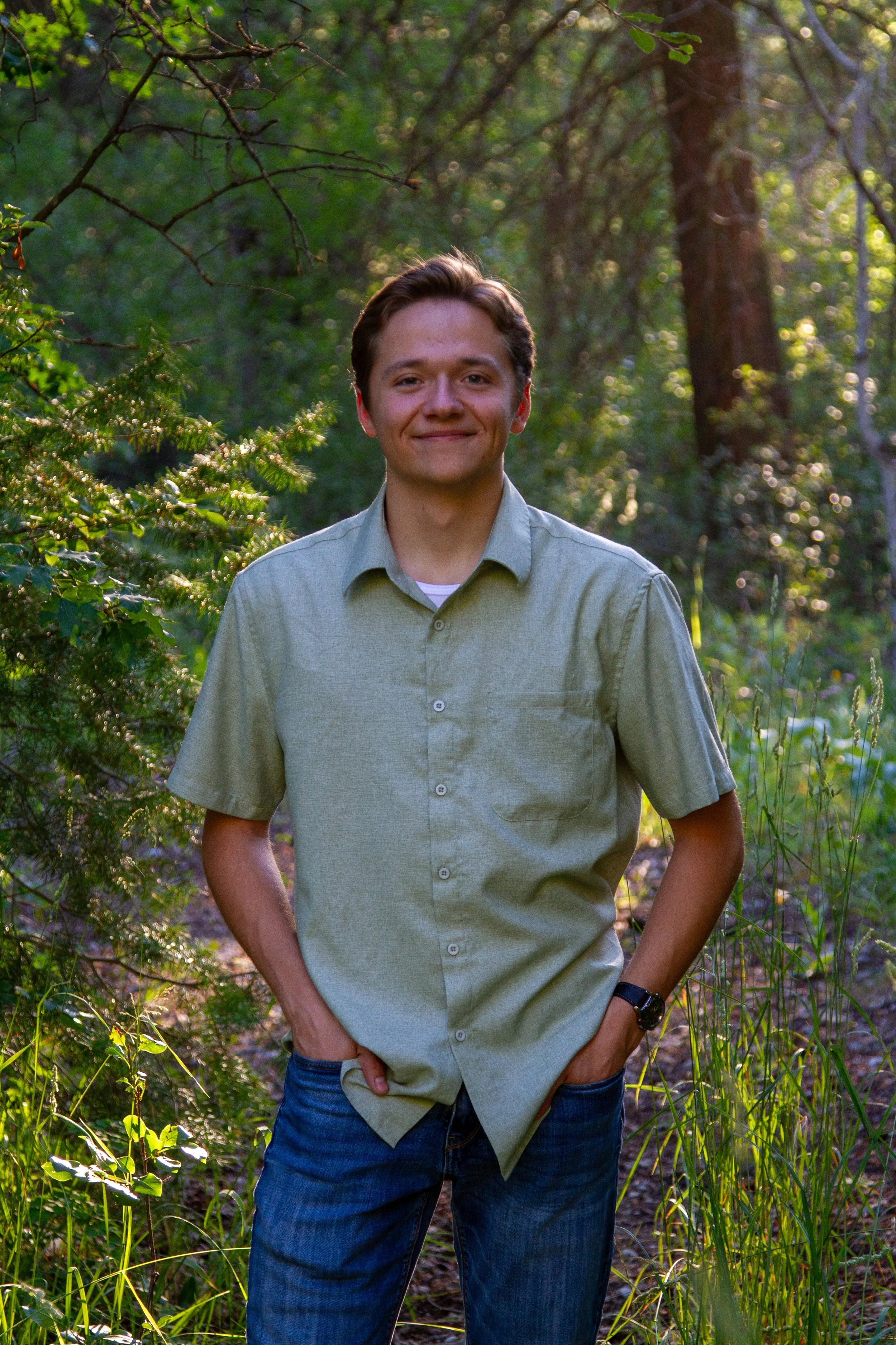 A young man with brown hair smiling and standing in a green forest during daylight, wearing a light green button-up shirt and blue jeans.