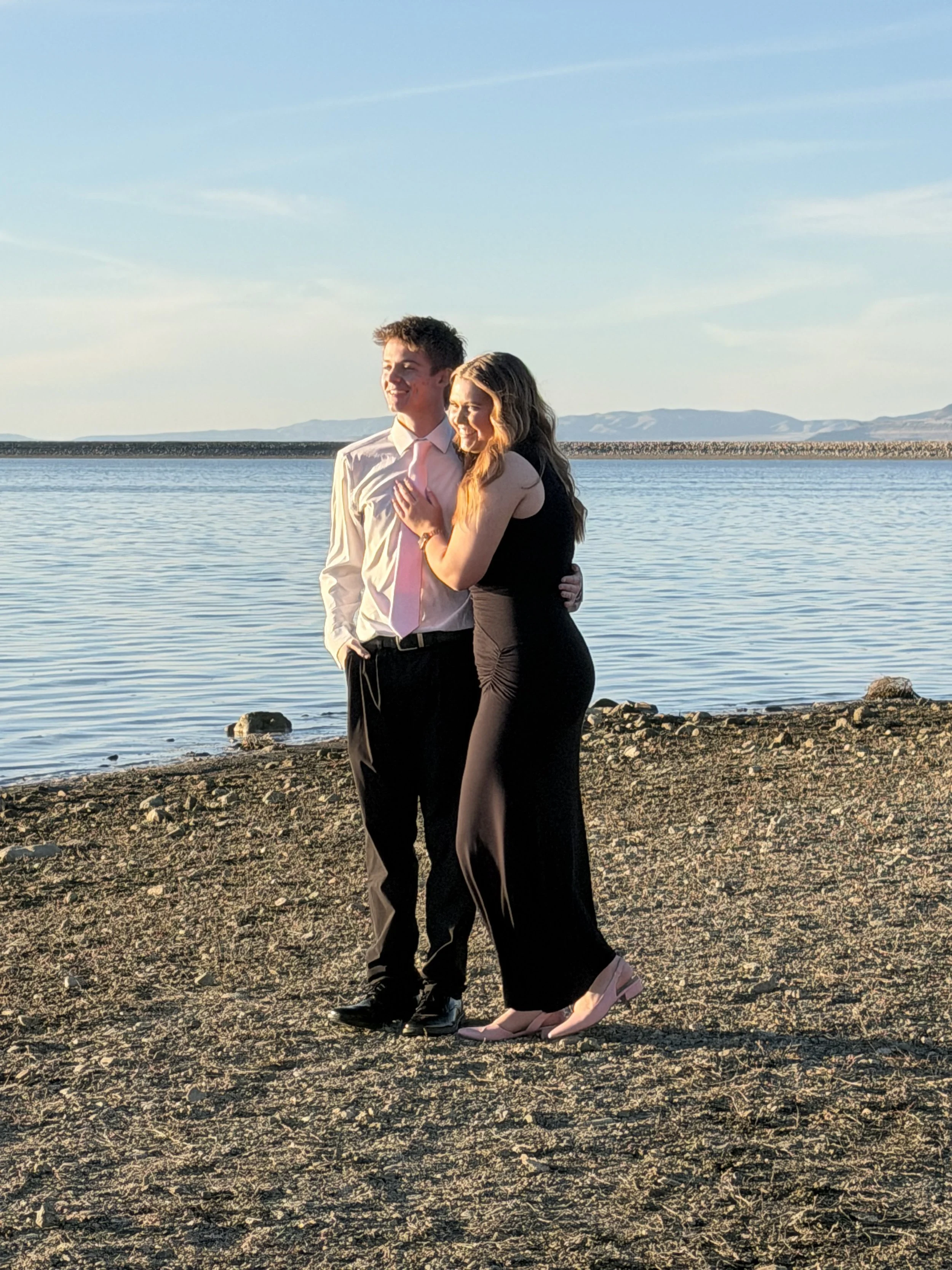 A young couple dressed in formal attire stands close together on a rocky beach near a body of water, smiling and enjoying each other's company during sunset.