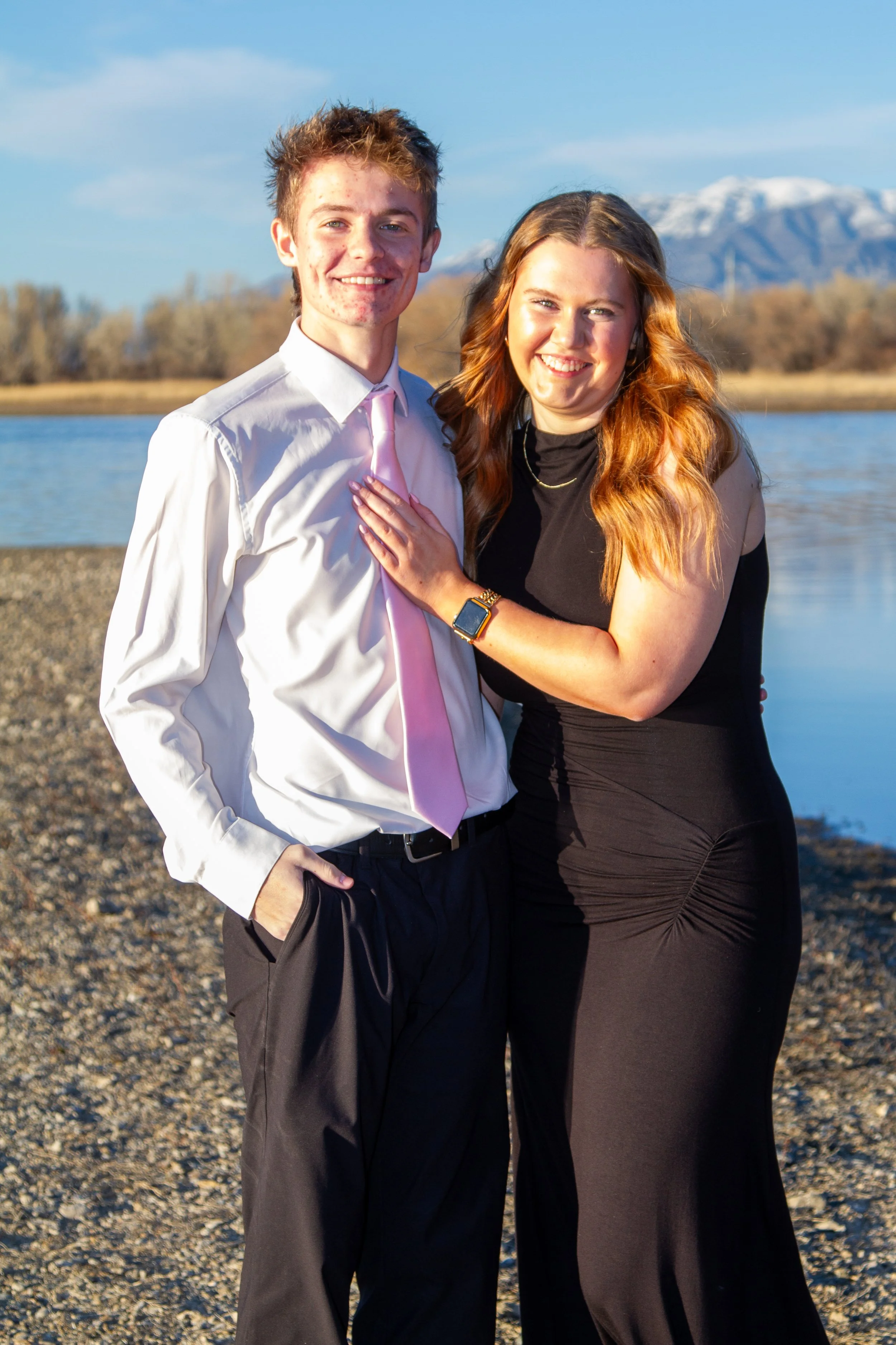 A young man in a white shirt and pink tie and a young woman in a black dress are standing close together outdoors near a river with a mountain in the background. They are smiling and the woman has her hand on the man's chest.