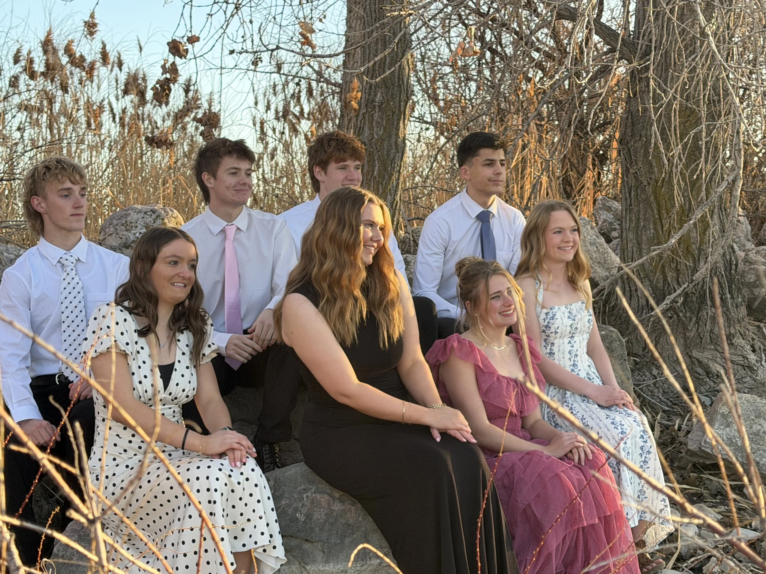 A group of nine young people dressed in semi-formal attire, sitting on rocks outdoors during sunset, smiling and posing for a photo.