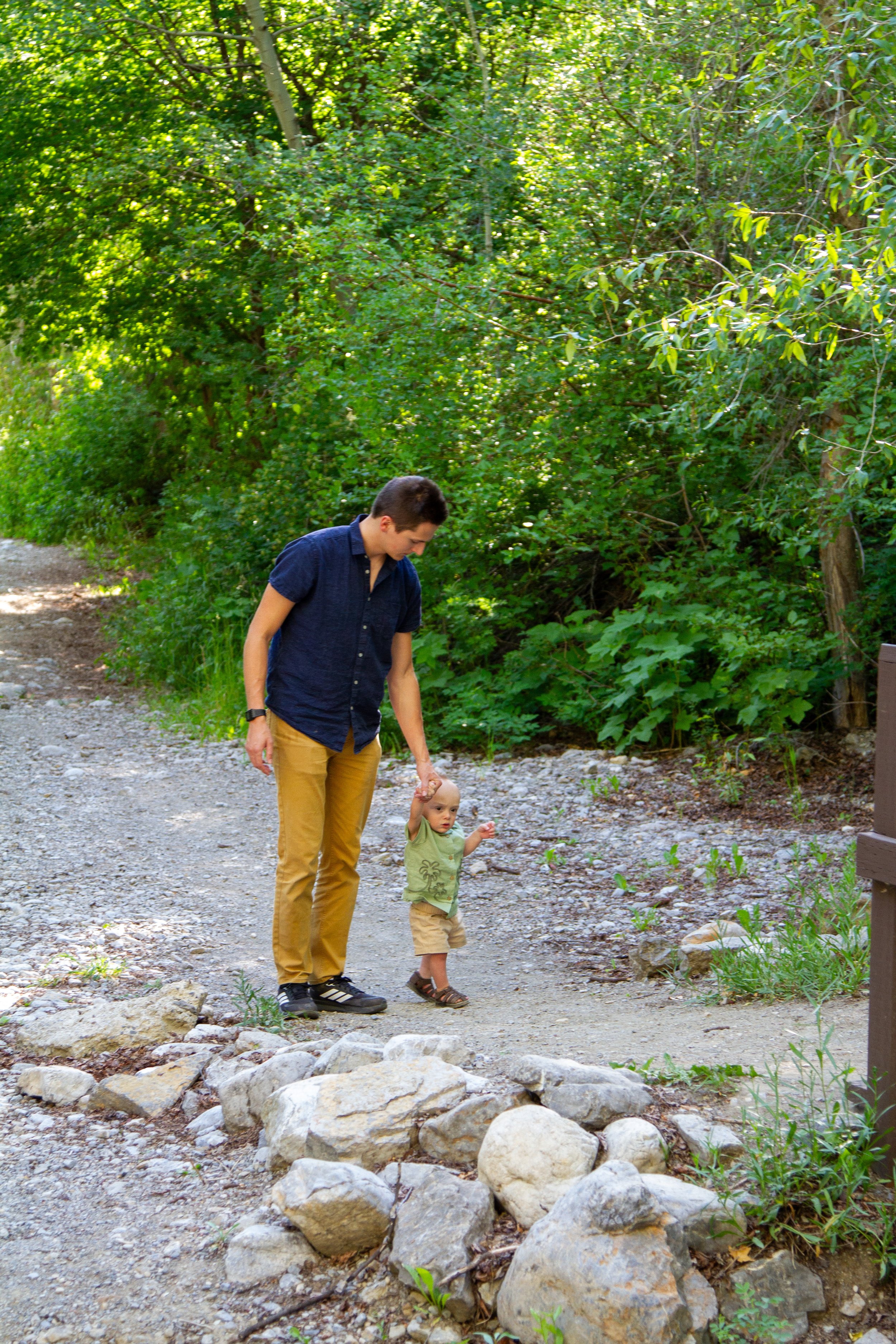 A young man in a dark blue shirt and khaki pants holding the hand of a small child wearing a green shirt and beige shorts on a rocky and gravelly trail surrounded by green trees and bushes.