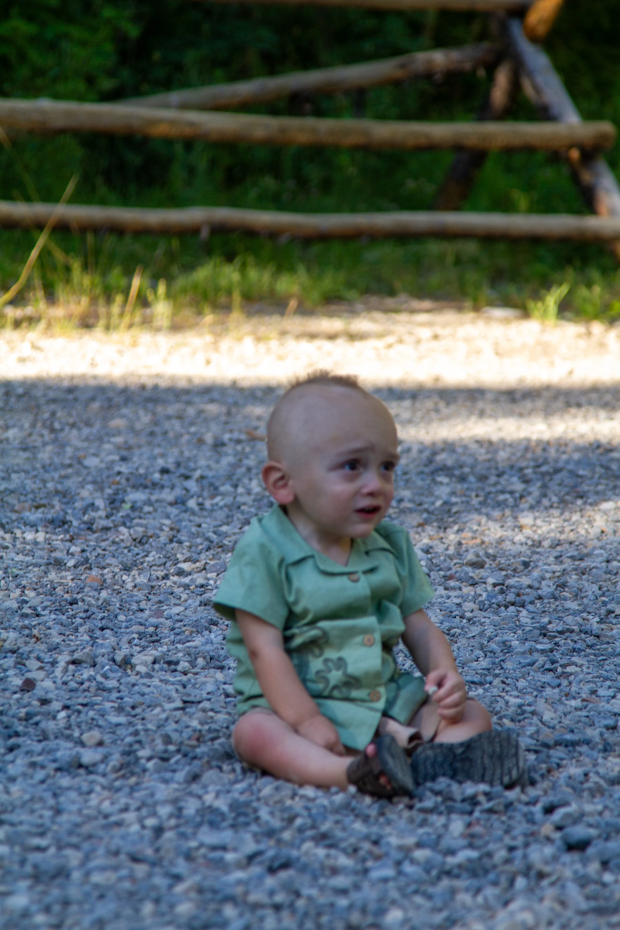 A young bald boy sitting on a gravel surface outdoors, with a wooden fence and green foliage in the background, wearing a green shirt and shorts, with a sad or distressed expression.