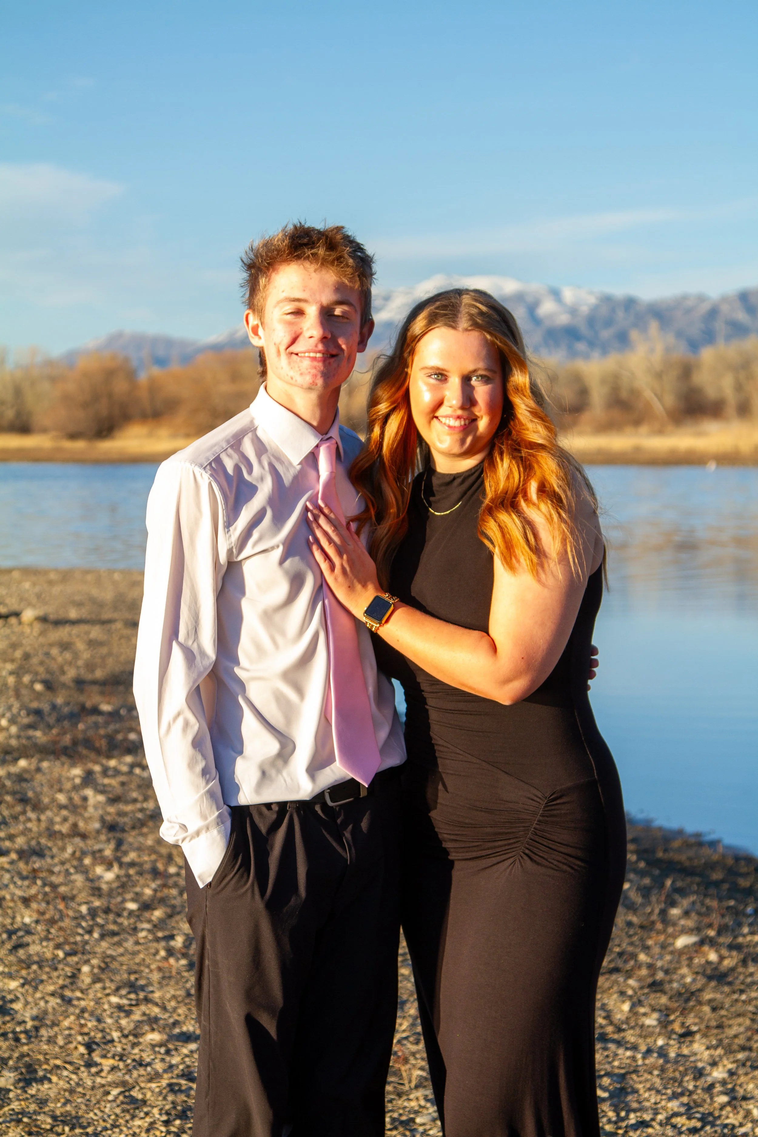 A young man in a white shirt and pink tie and a young woman in a black dress standing together by the water on a rocky beach with mountains in the background during sunset.