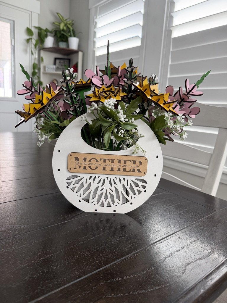 A decorative flower arrangement in a white, round, cut-out vase with a wooden sign that says "Mother" and an inscription 'You're the best person in the world.' The arrangement includes pink, yellow, and purple paper flowers with green leaves, placed 