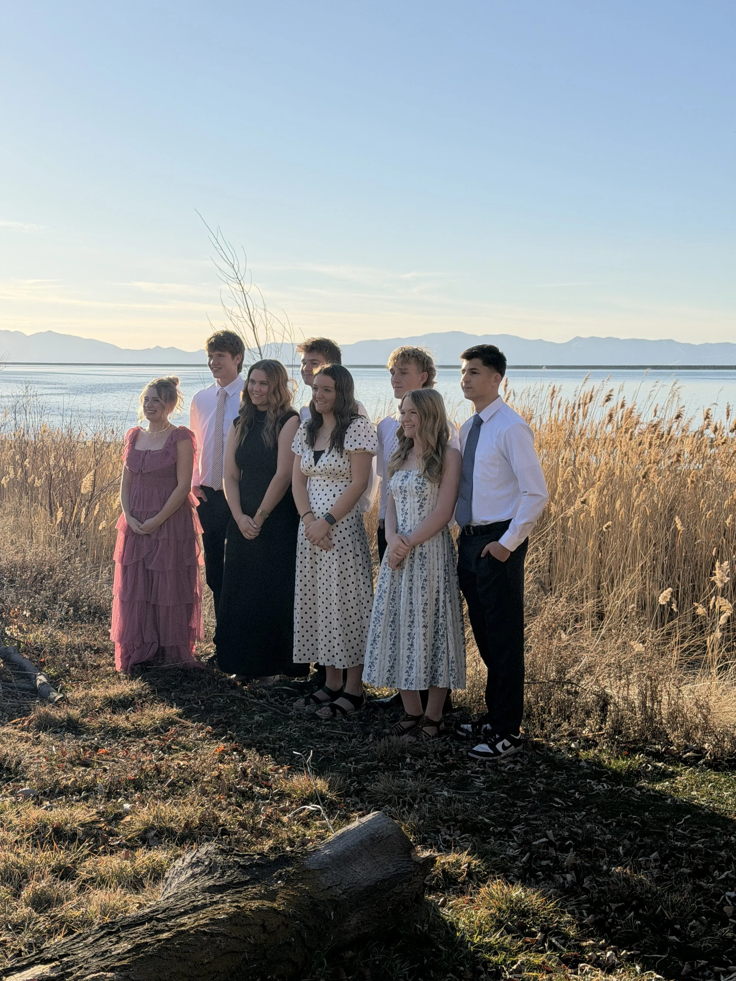 A group of eight young people posing outdoors by a body of water with mountains in the background, during sunset. The group includes six women and two men, dressed in casual and semi-formal attire, standing on grass and dried plants.