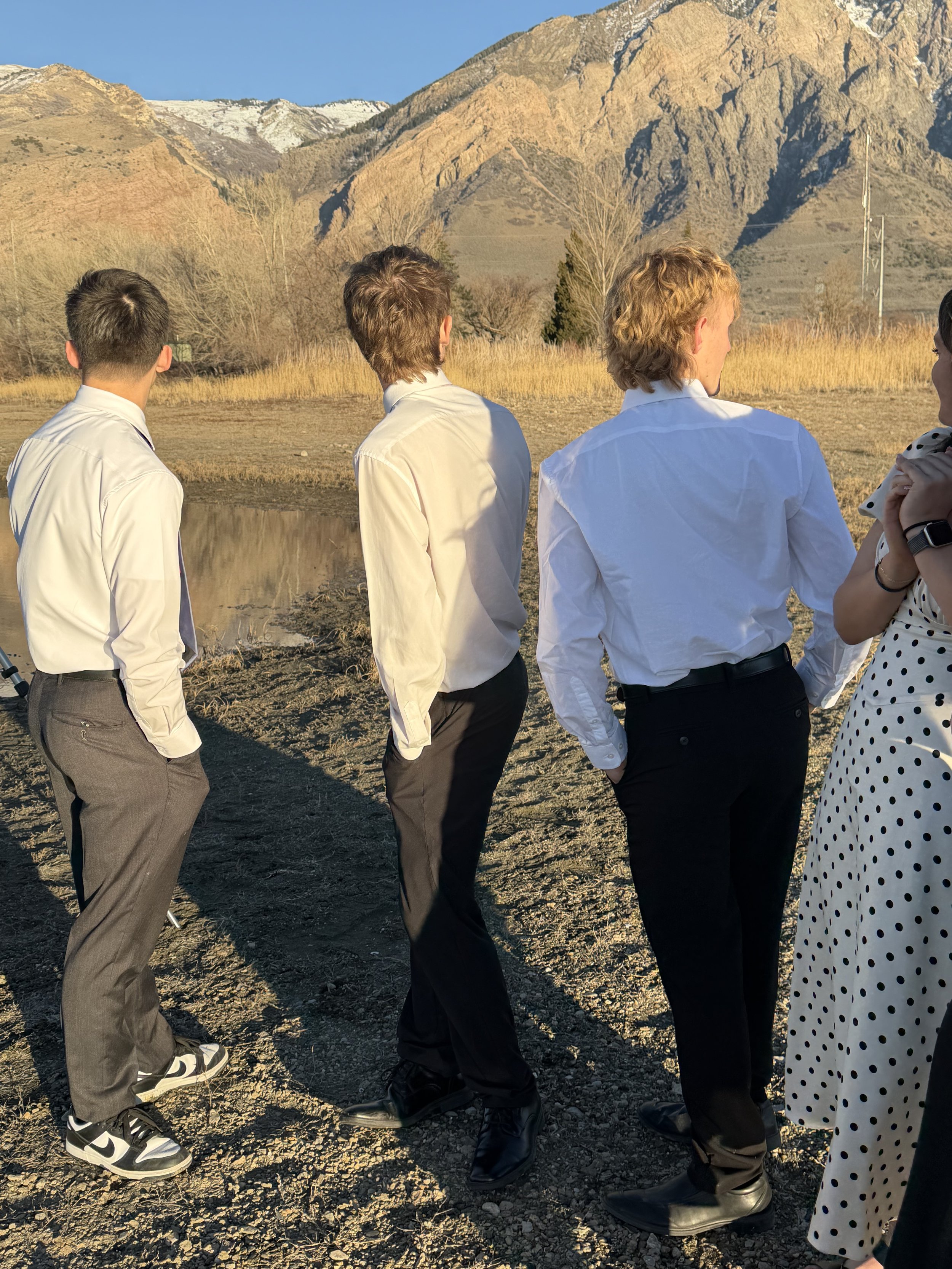 Group of four people in formal attire standing outdoors on rocky ground, with mountains and dry grass in the background during sunset.