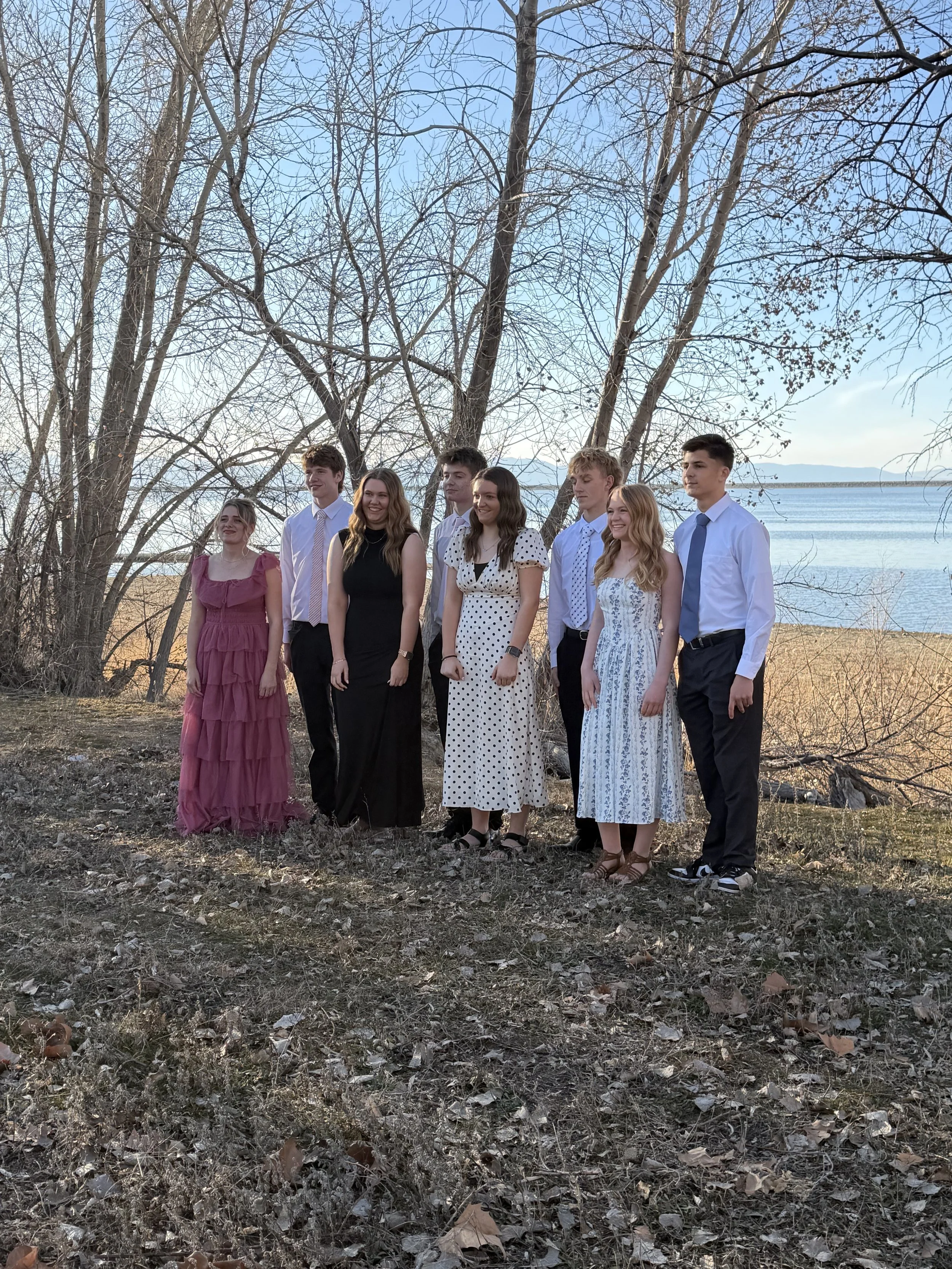 Group of eight young people dressed in semi-formal attire standing outdoors near a body of water and leafless trees on a clear day.