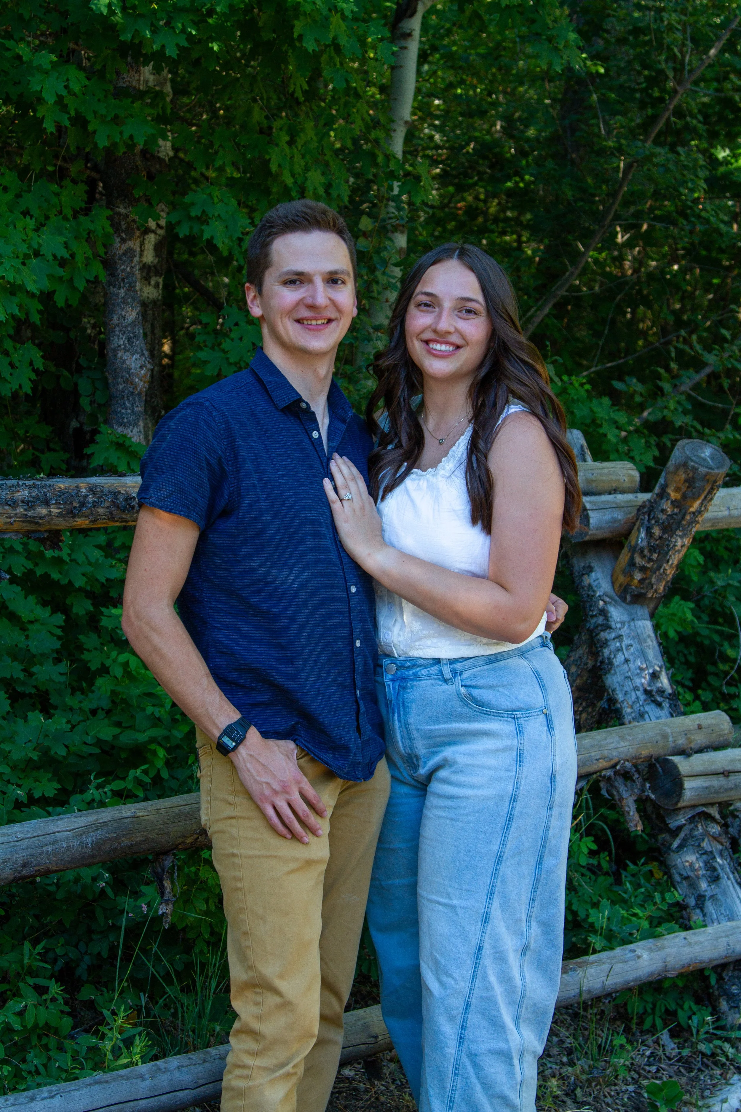A young man wearing a blue shirt and tan pants standing next to a young woman in a white top and light blue jeans, posing outdoors with green trees and a wooden fence in the background.