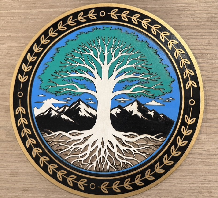 Decorative round artwork featuring a large white tree with roots, mountains, and clouds inside a black and gold border with leaf patterns, on a wooden surface.