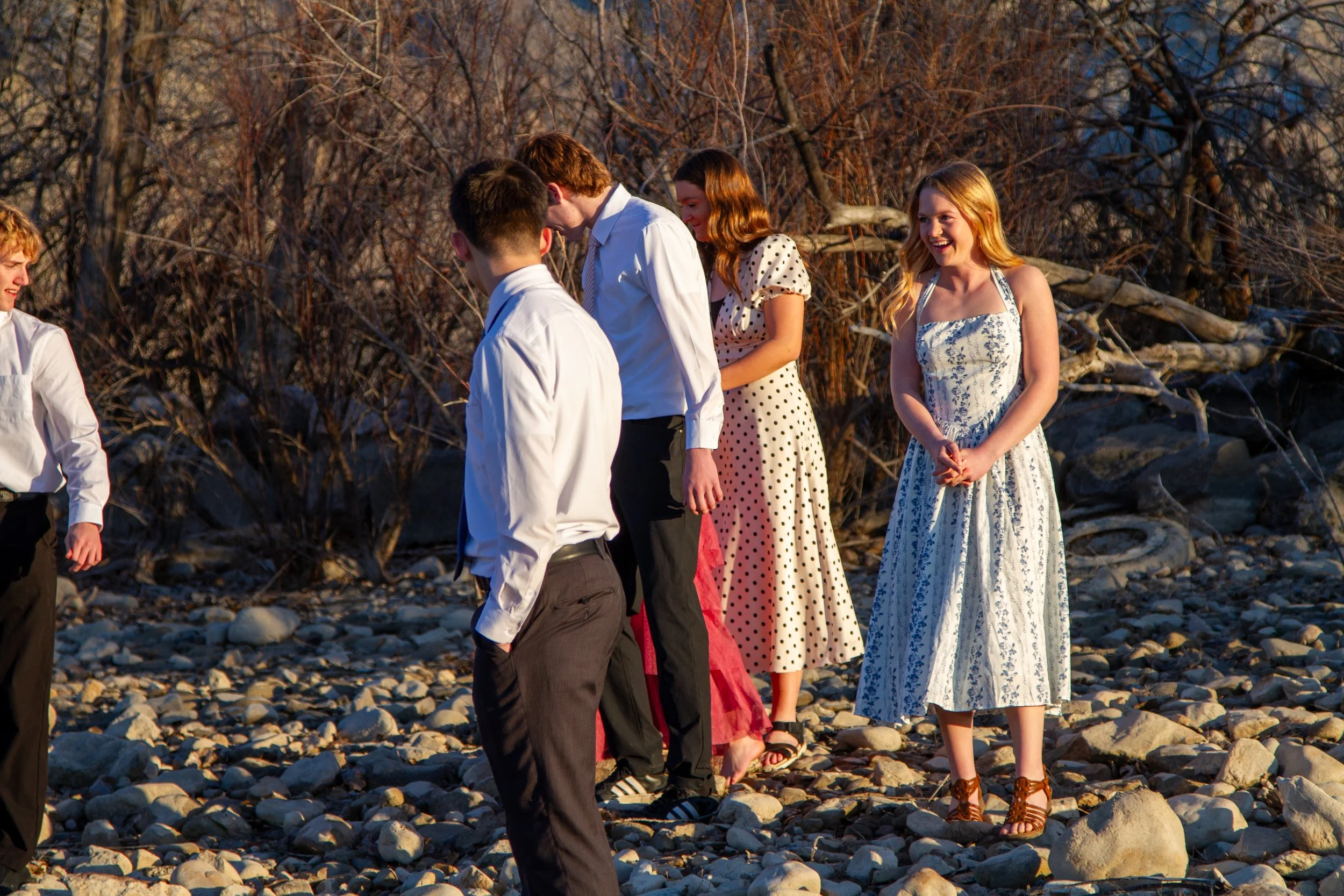Group of young people in casual and semi-formal clothes standing on a rocky beach with bare trees in the background during sunset