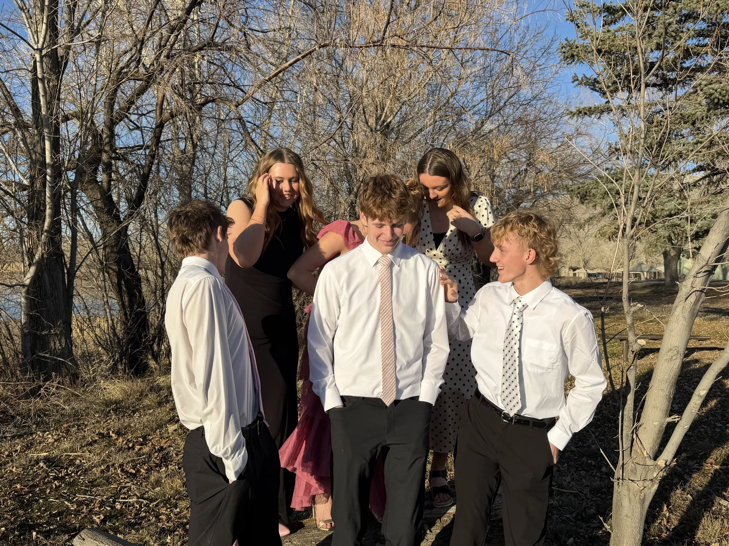 Group of five young adults standing outdoors among leafless trees, dressed in semi-formal attire, engaging and smiling at each other on a sunny day.
