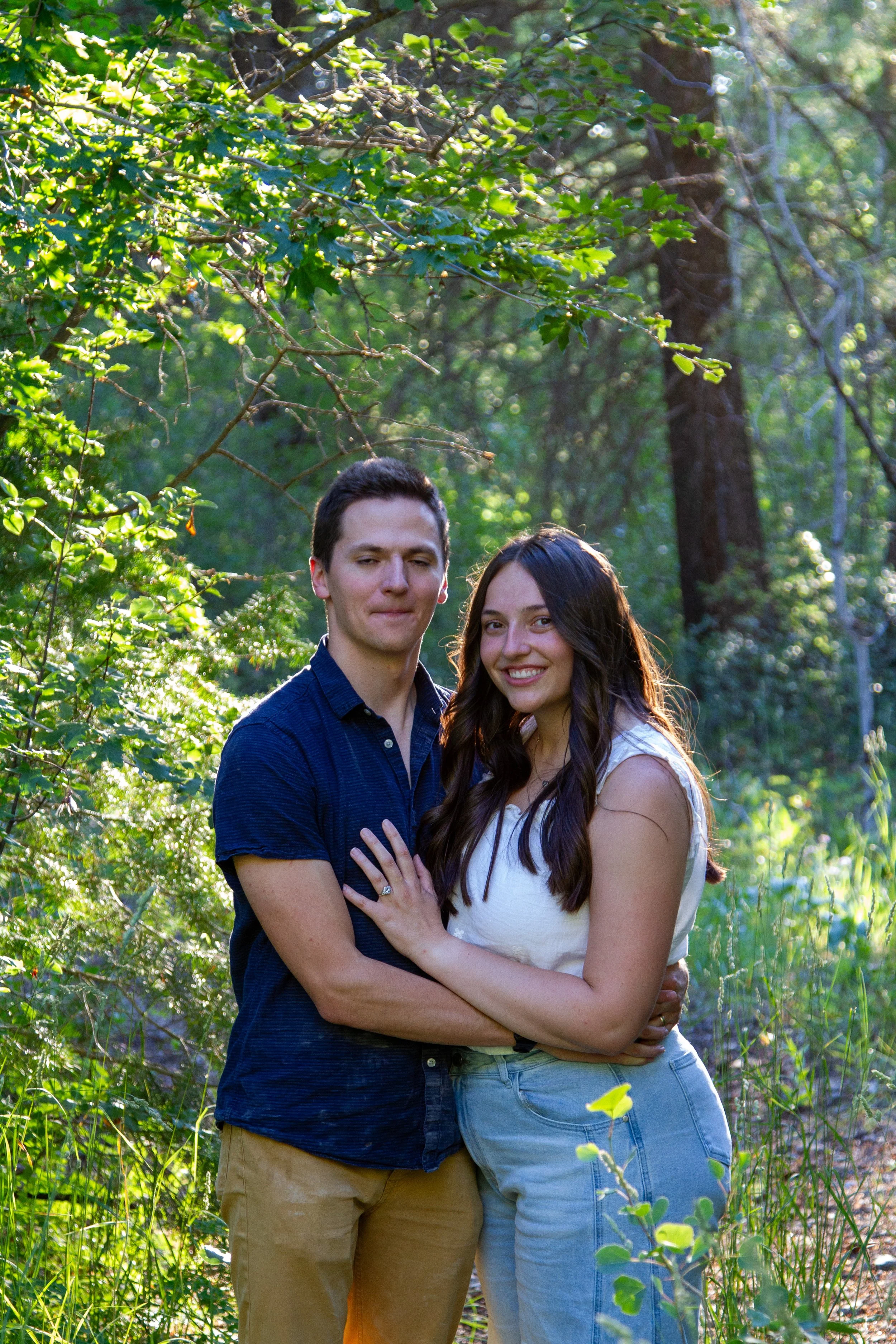 A young couple standing close together outdoors among green trees and plants, smiling with arms around each other.