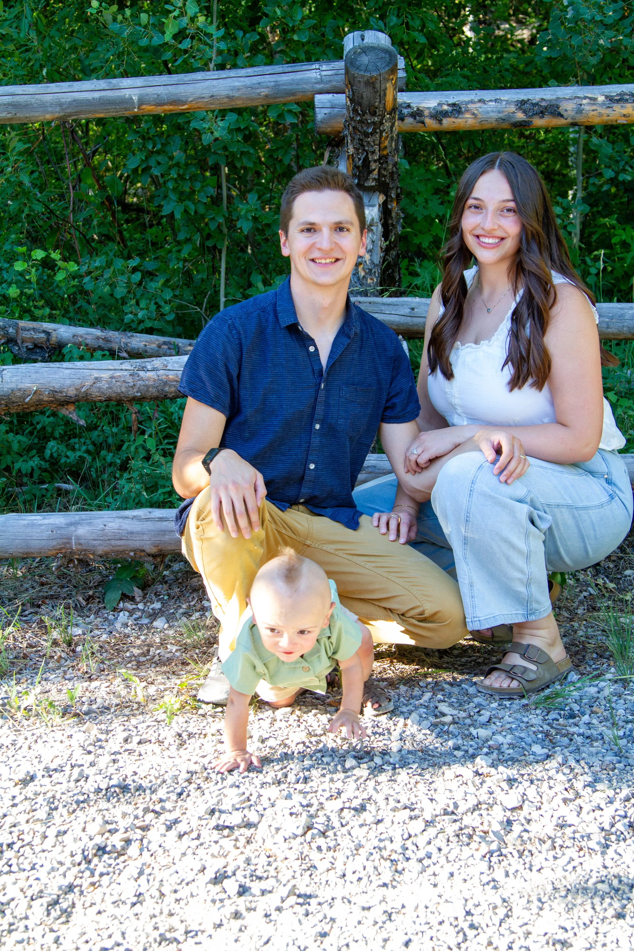 A smiling young couple sitting on a wooden fence outdoors with two children, one crawling on the ground and another sitting, surrounded by greenery.
