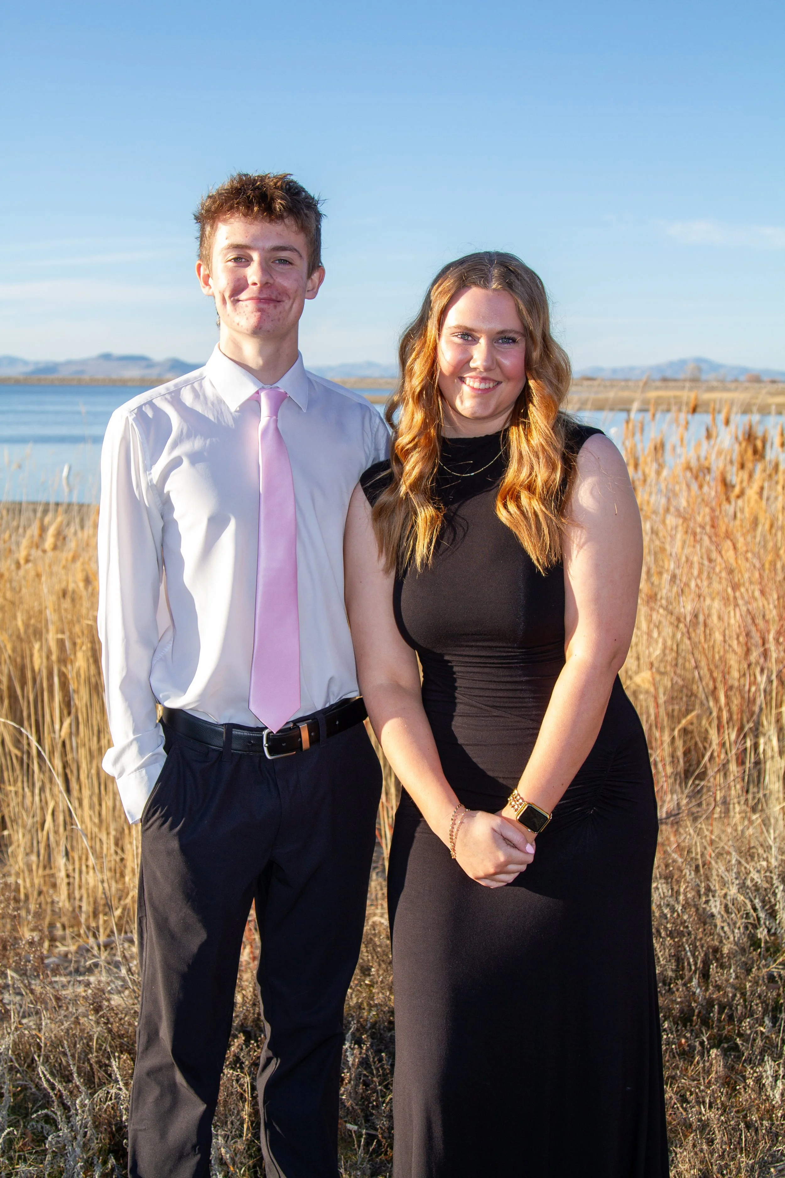 A young man and woman standing outdoors near a body of water with mountains in the background, smiling at the camera. The man is wearing a white shirt and pink tie, and the woman is in a black dress.