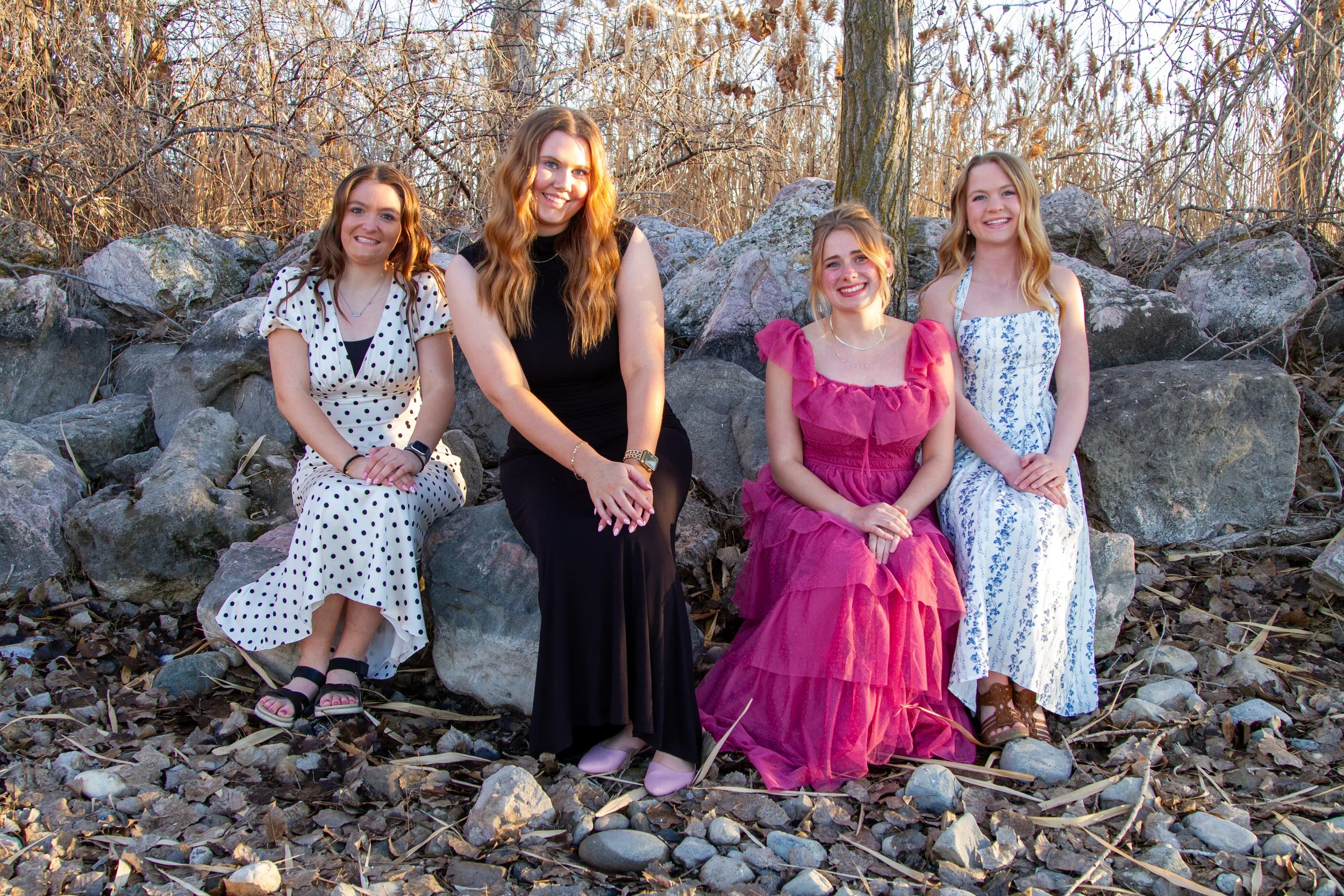 Four women sitting on rocks outdoors during daylight, dressed in colorful dresses, with leafless trees and rocks in the background.