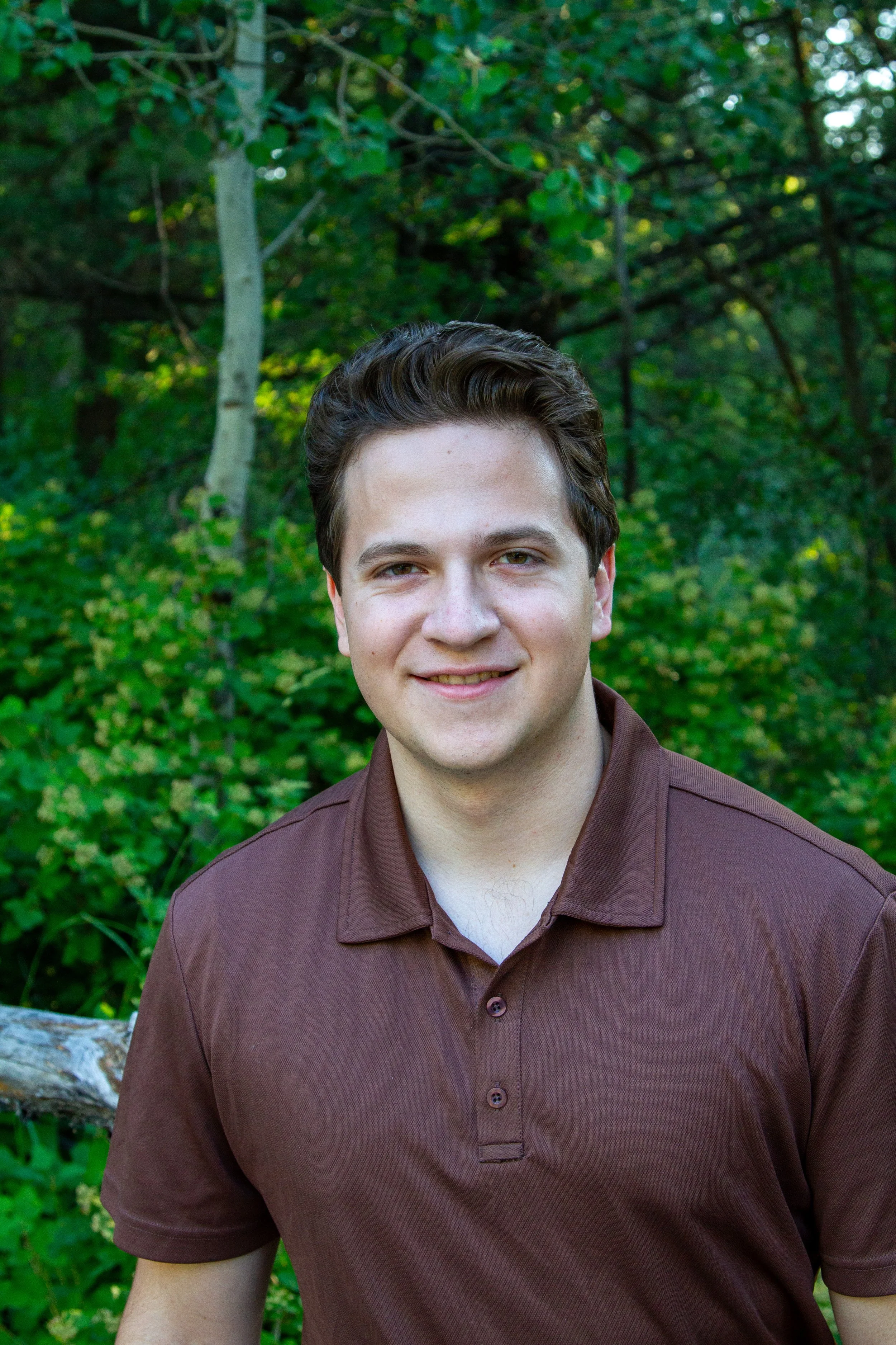 Young man smiling outdoors among green trees and foliage.