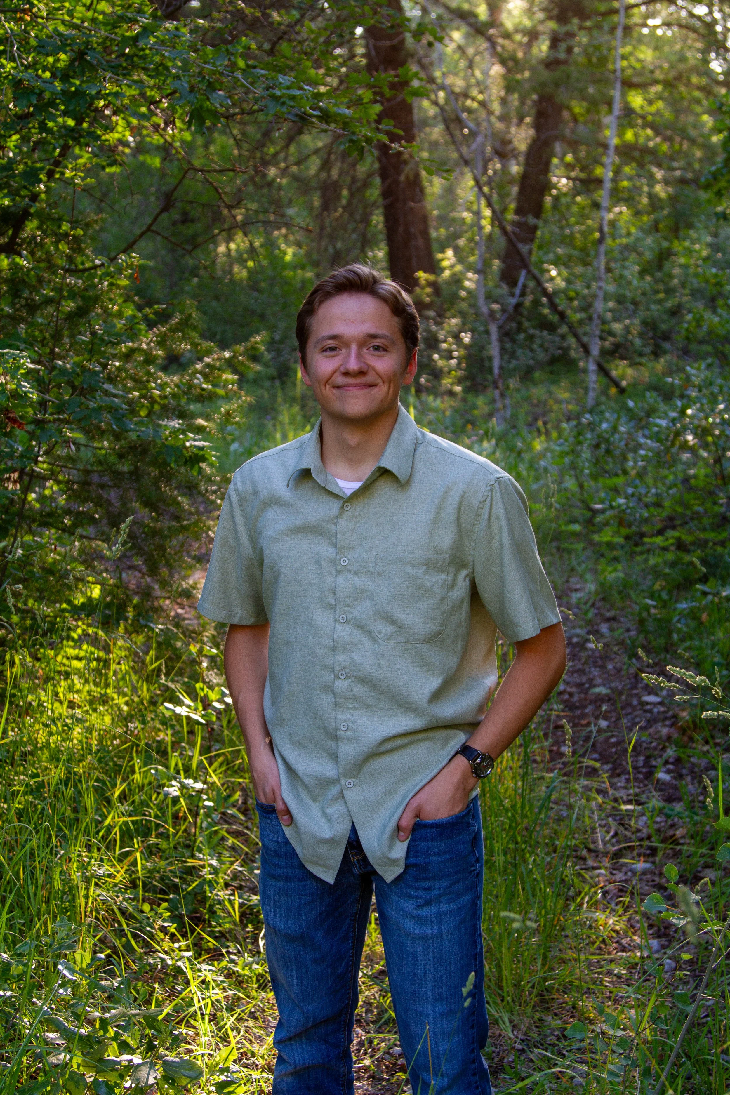 A young man with short brown hair smiling outdoors in a green, wooded area, wearing a light green button-up shirt and blue jeans.