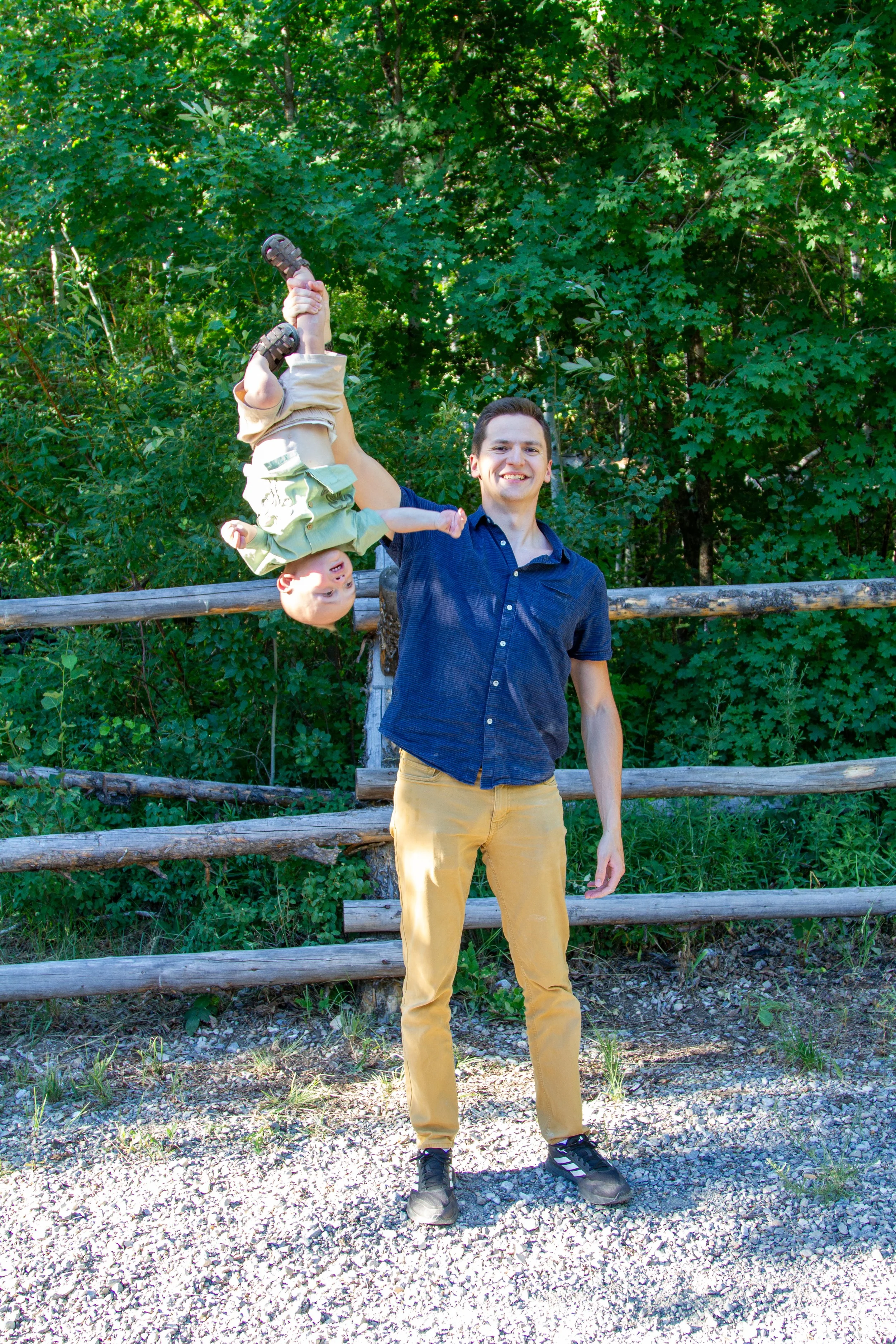 A man standing outdoors on a gravel path, holding a smiling baby with fingers pointing upward, while the baby is hanging upside down by the man's hand, against a background of lush green trees and a wooden fence.