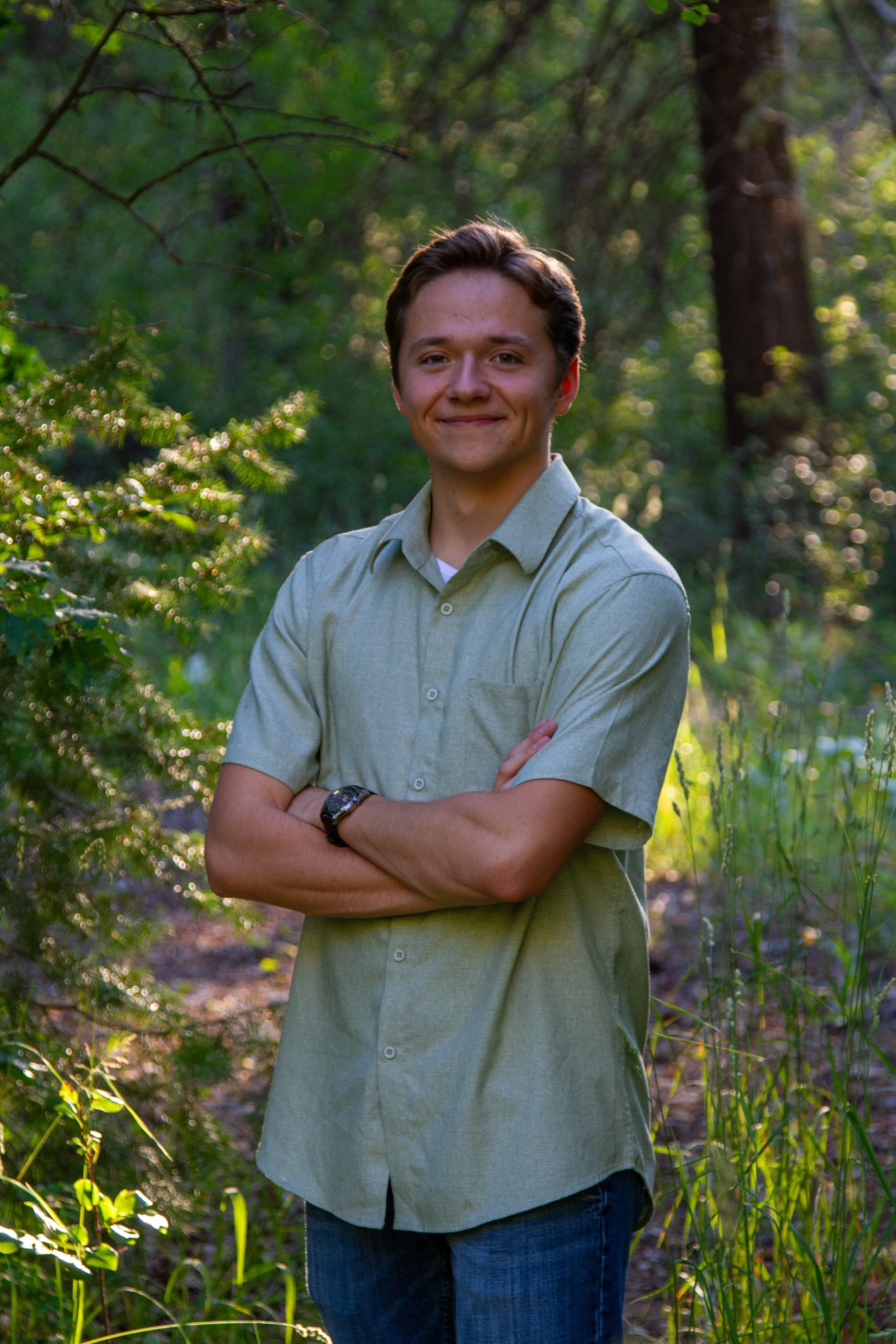 A smiling young man with light skin and brown hair, wearing a light green button-up shirt and jeans, stands with arms crossed in a lush green forest with sunlight filtering through trees in the background.