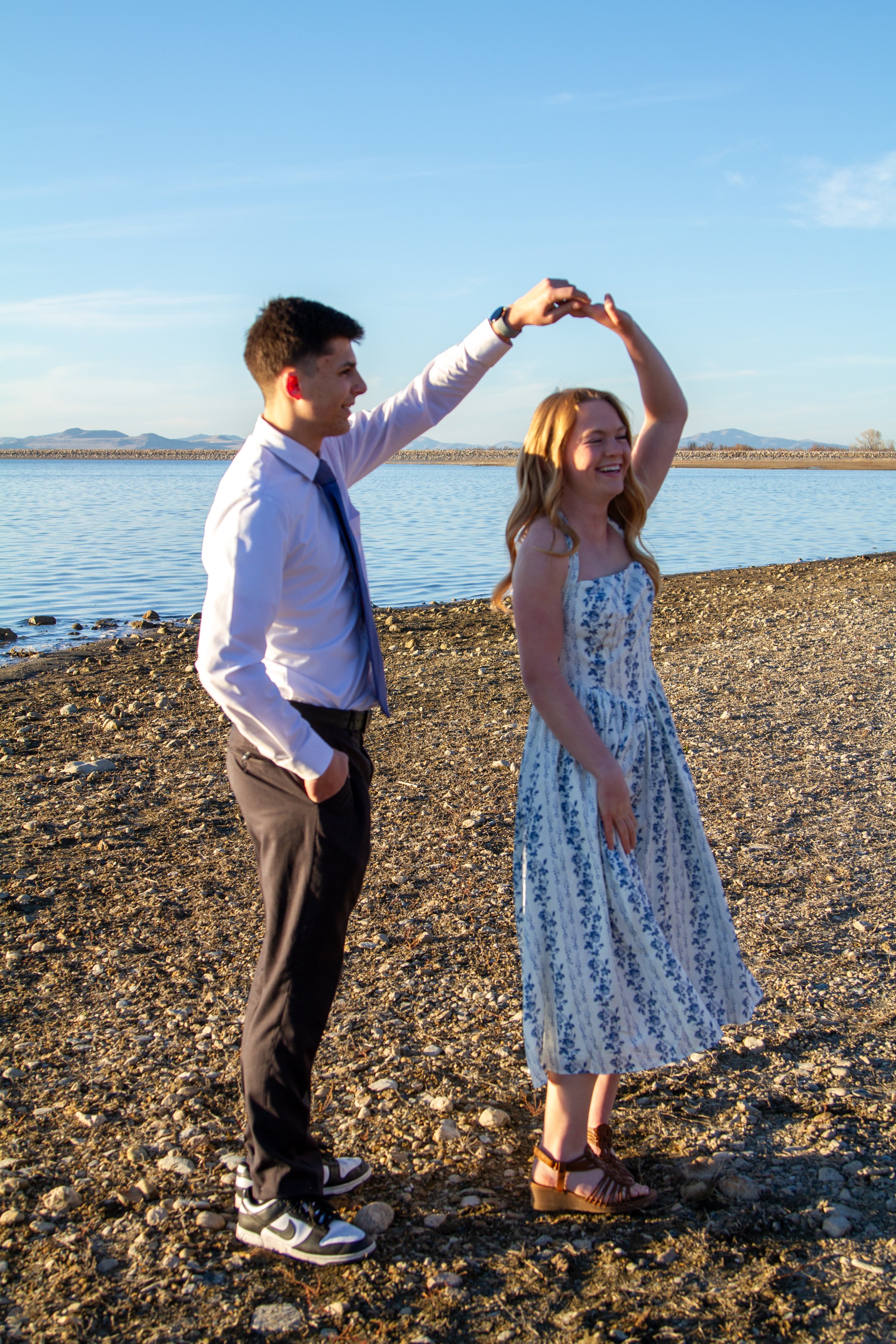 A young man and woman are happily dancing and holding hands by the lakeside on a sunny day, with a clear blue sky and mountains in the background.