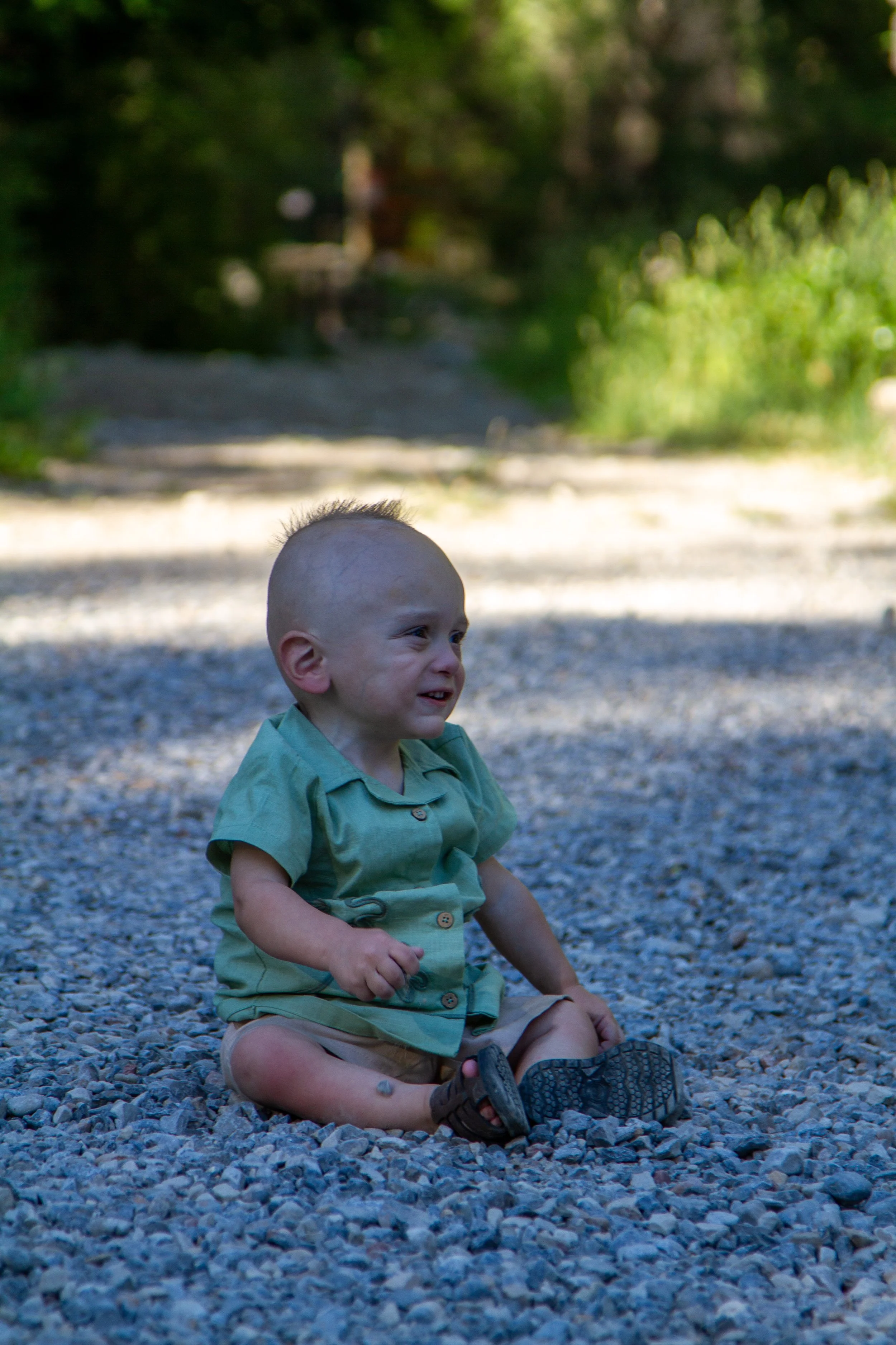 A young boy with a mohawk haircut sitting on a gravel surface outdoors, smiling and wearing a green shirt and shorts, with trees and greenery in the background.