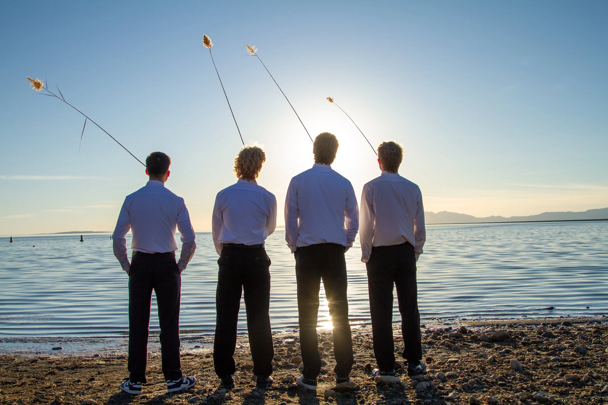 Four men in dress shirts and pants standing on a rocky beach facing the water at sunset, with three fishing rods cast into the water.