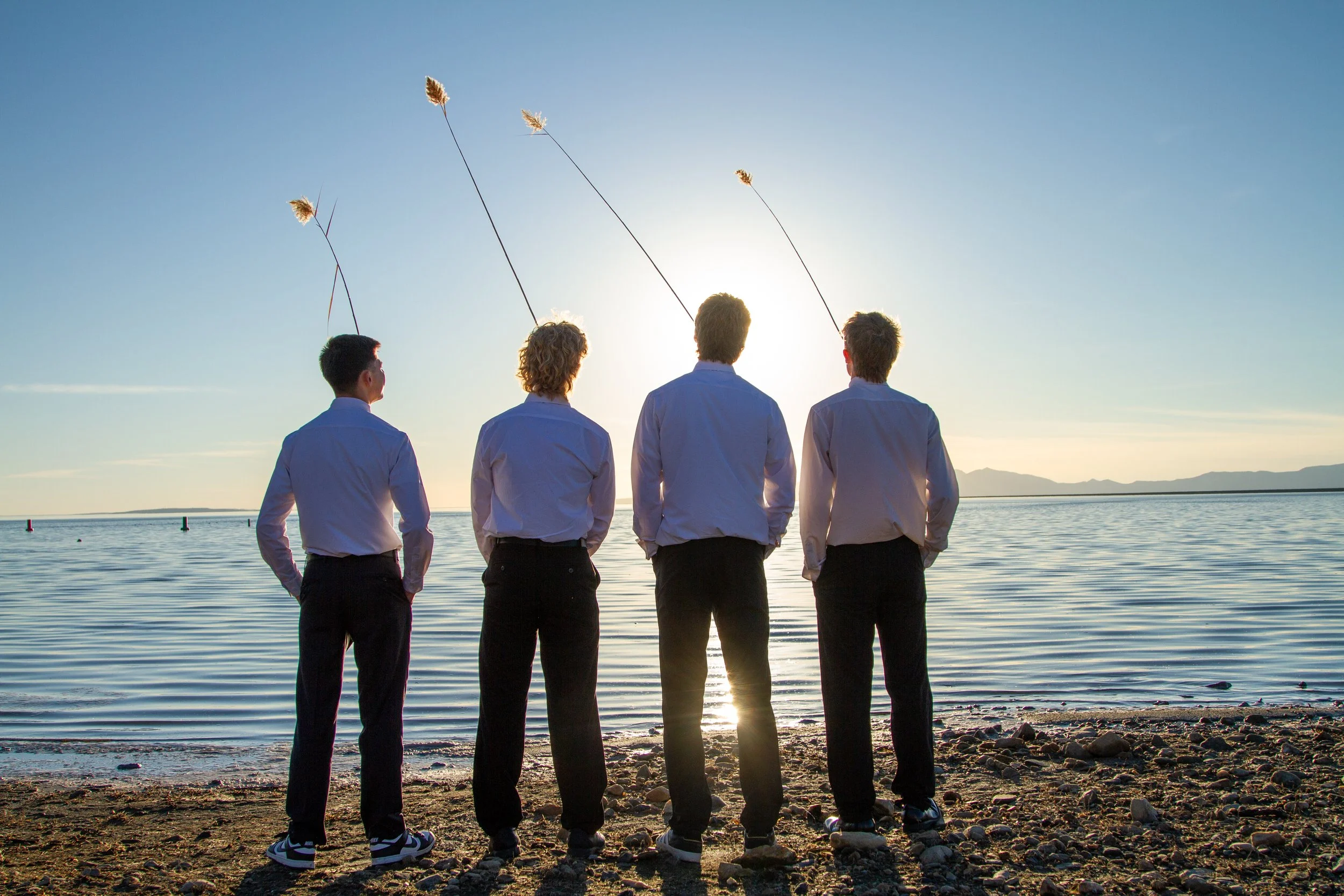 Four people stand on a rocky shoreline facing the water at sunset, holding long sticks with items at the end, resembling fishing or kite flying.